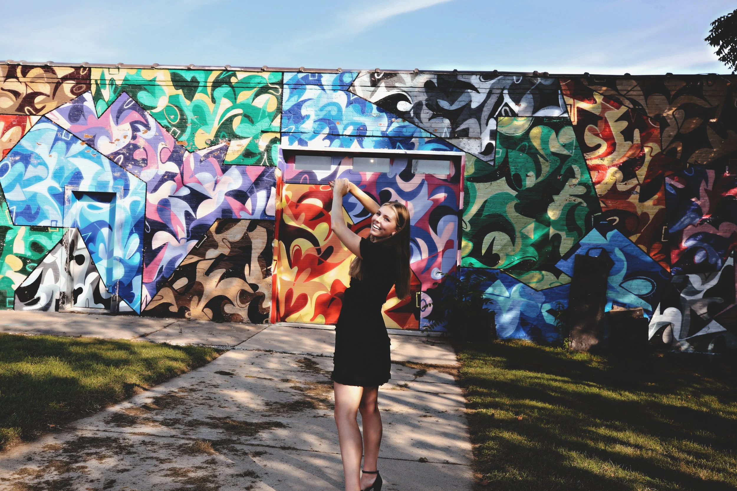 A woman in a black dress and high heels standing in front of a colorful graffiti mural, smiling and posing with her arms raised.