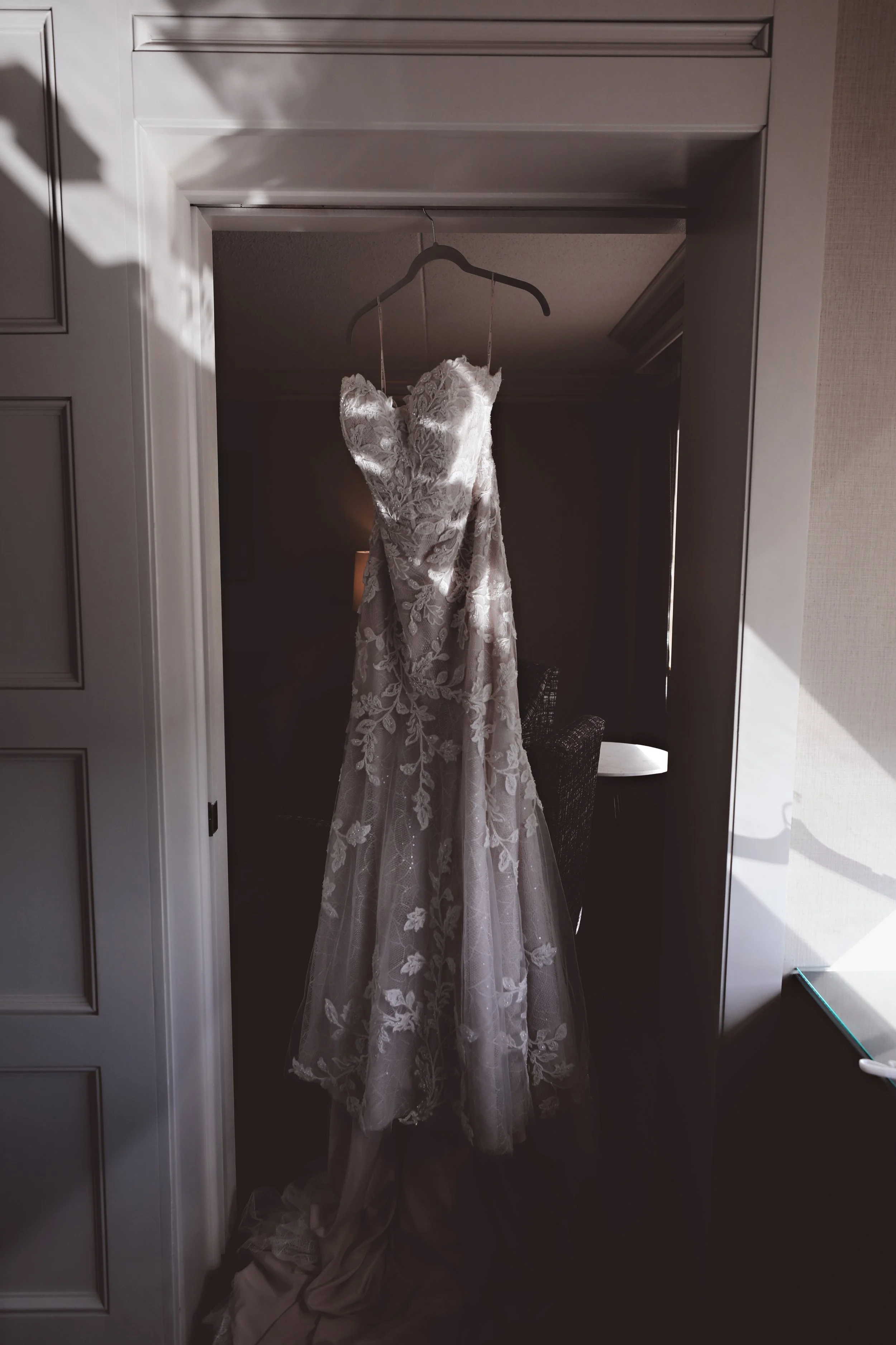 A wedding dress hanging on a hanger inside a doorway with sunlight casting shadows.
