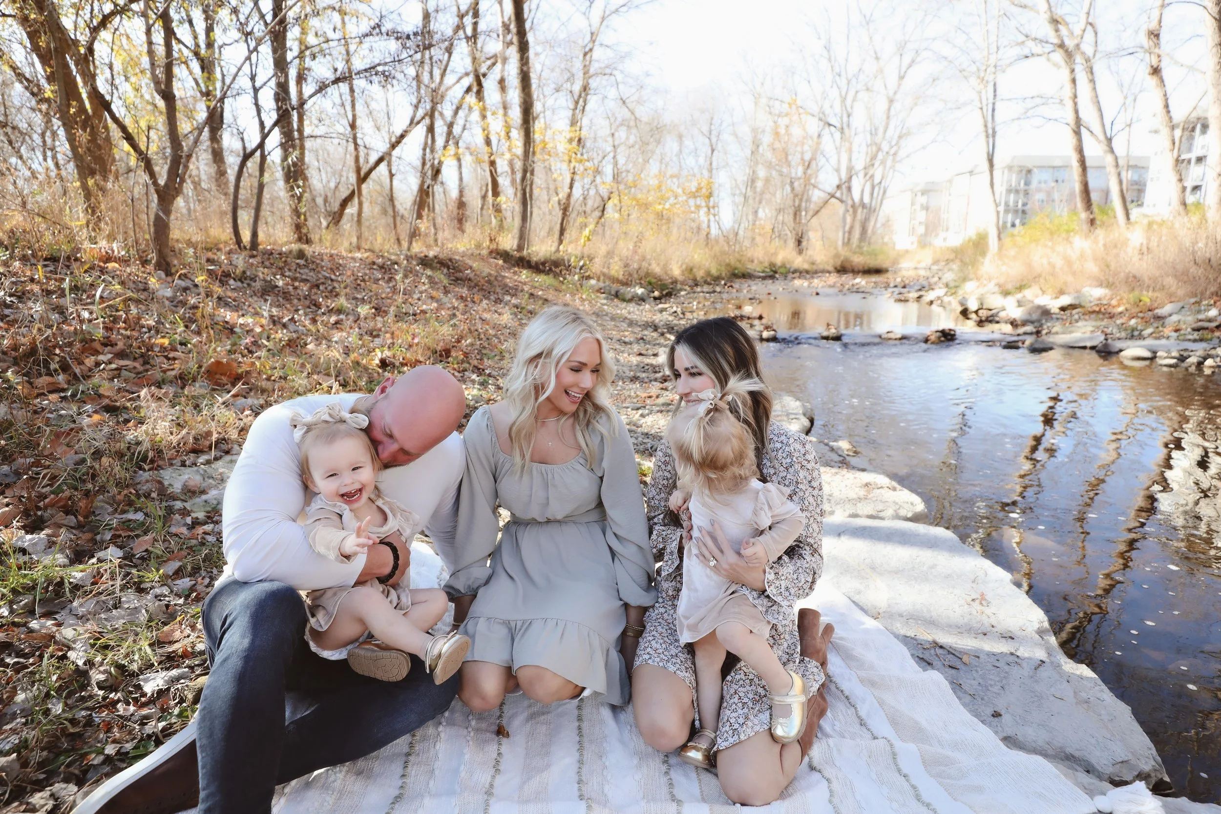 Family of four enjoying a picnic by a creek in a wooded area on a sunny day.
