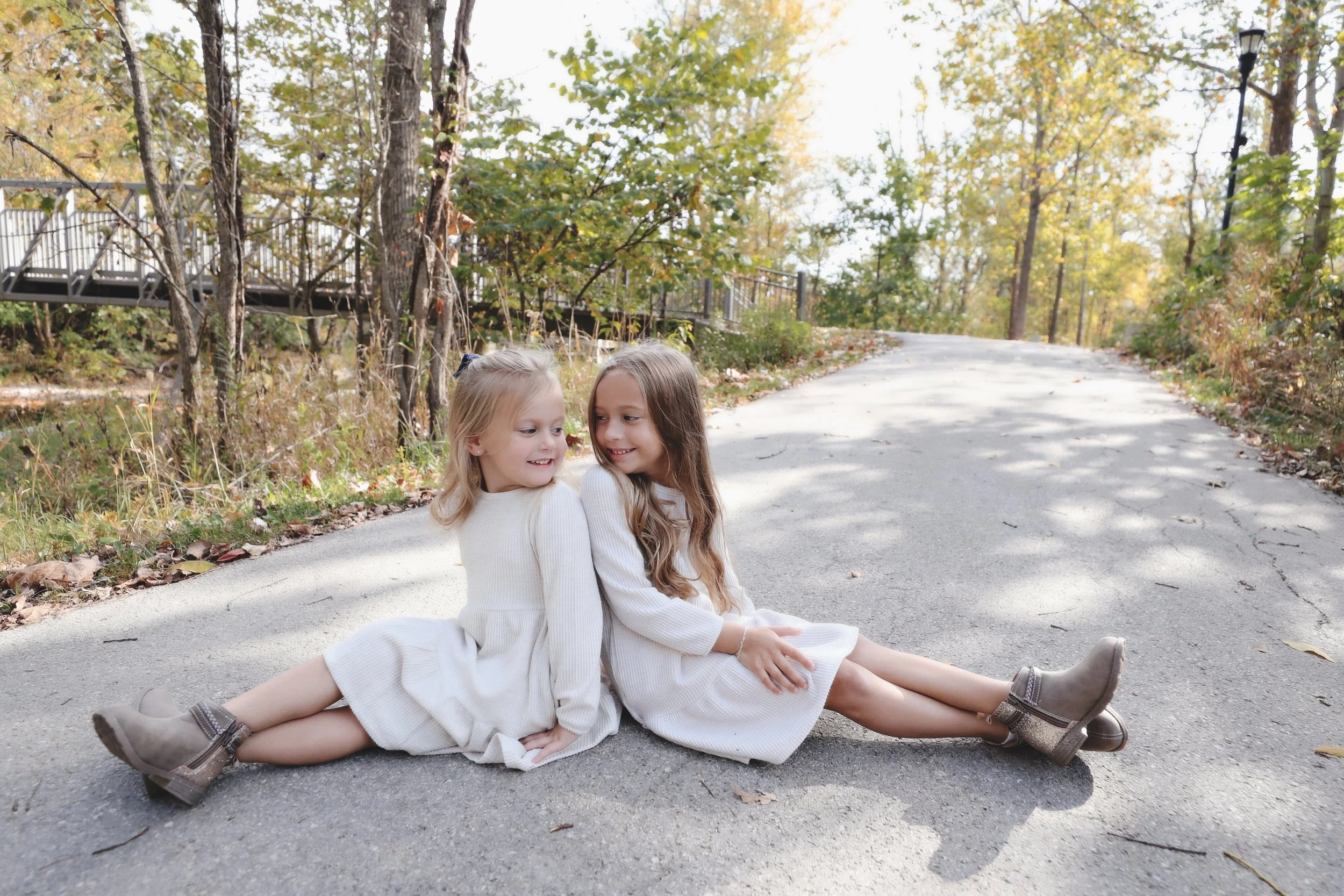 Two young girls in white dresses and boots sitting on a paved path in a woodland area, smiling at each other with trees and a railing in the background during autumn.