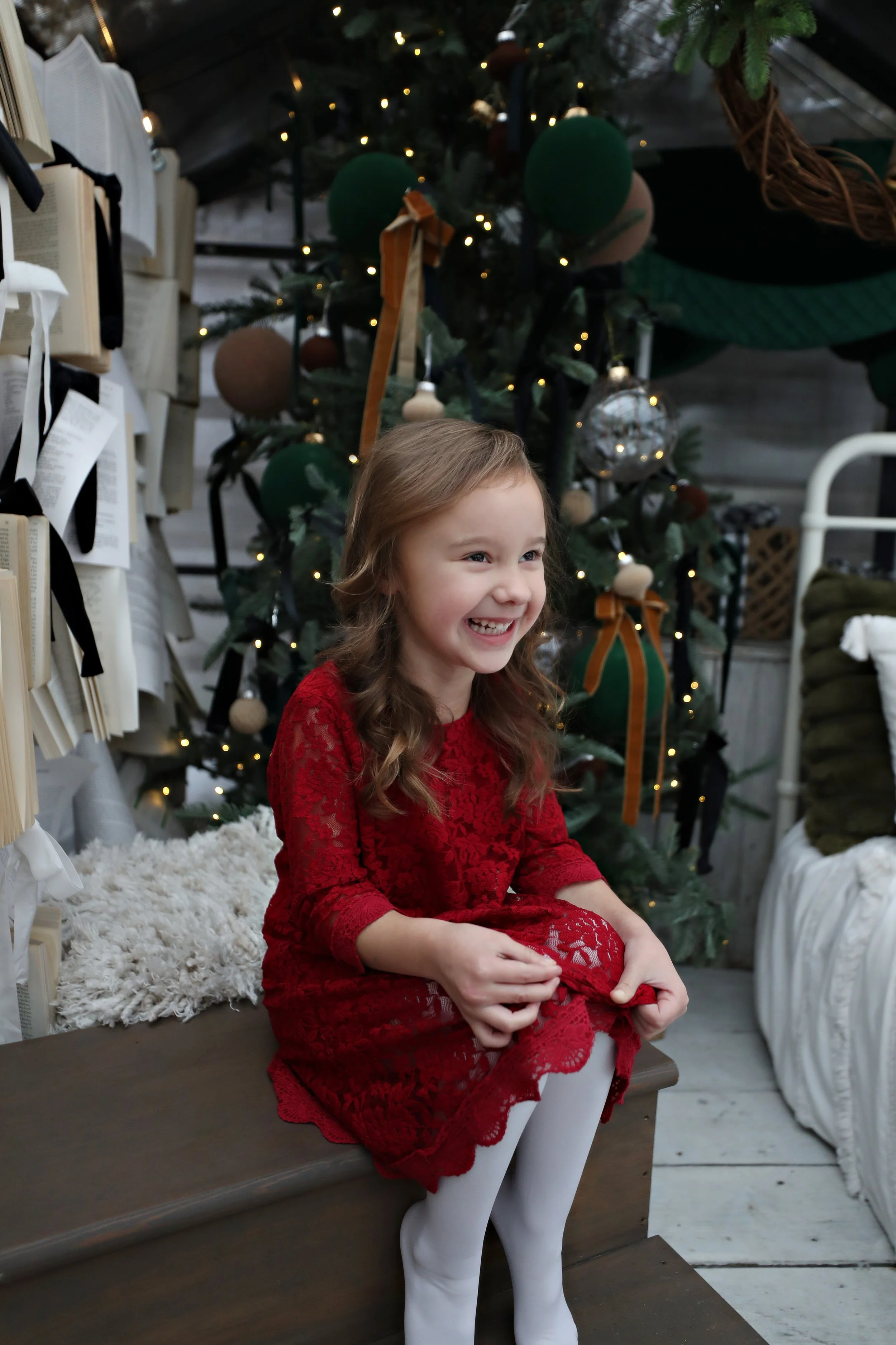 A young girl in a red lace dress sitting on a wooden bench in front of a decorated Christmas tree, smiling and happy.