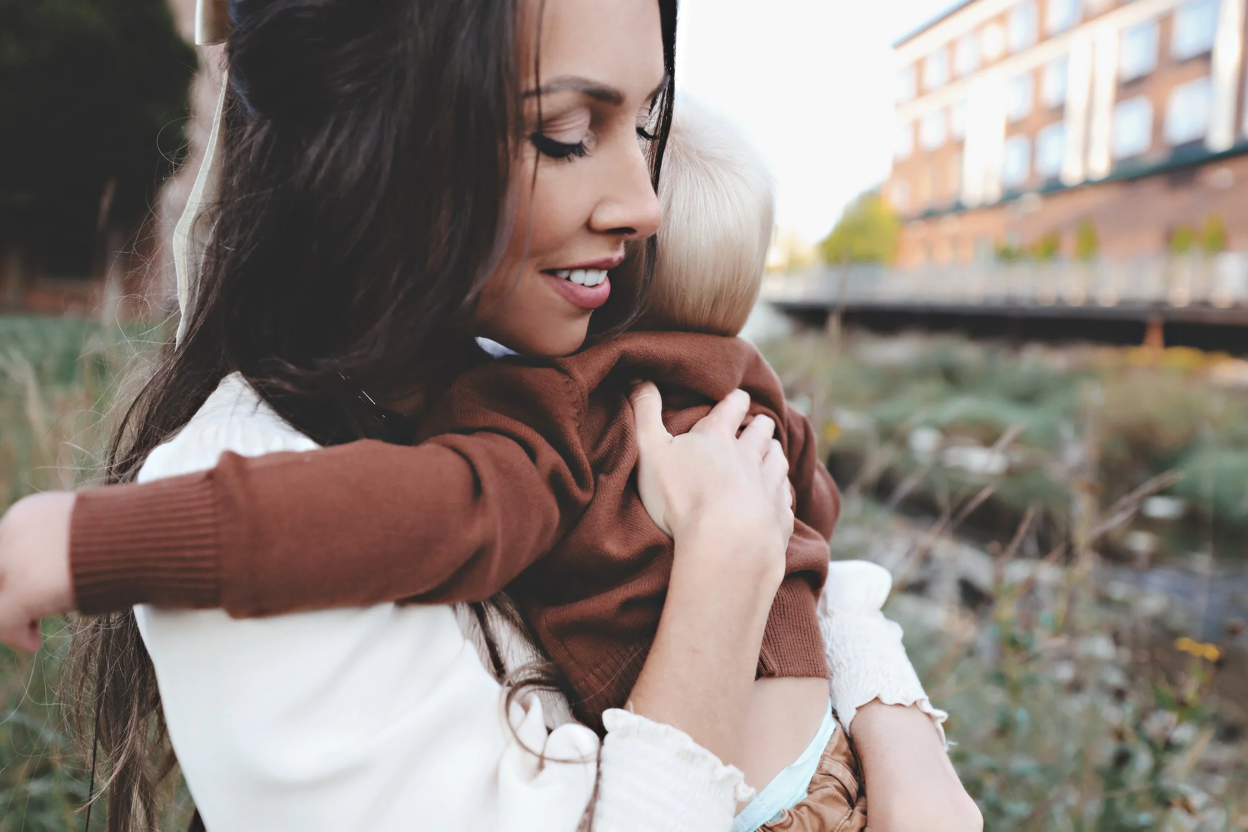 Woman hugging a young child outdoors near a river and buildings