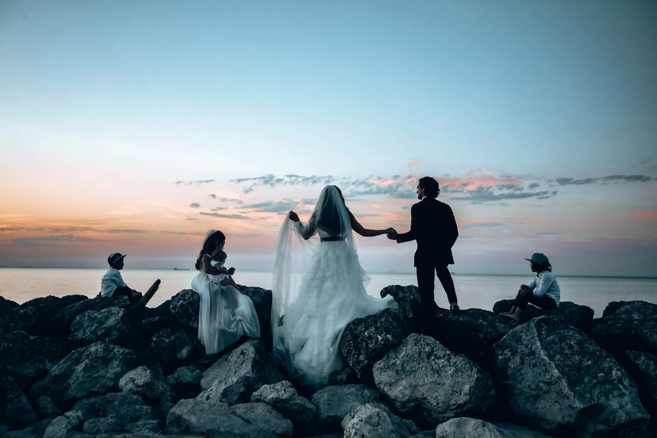 A wedding couple standing on rocks by the water during sunset, with children sitting nearby, some using devices, and the bride and groom holding hands.