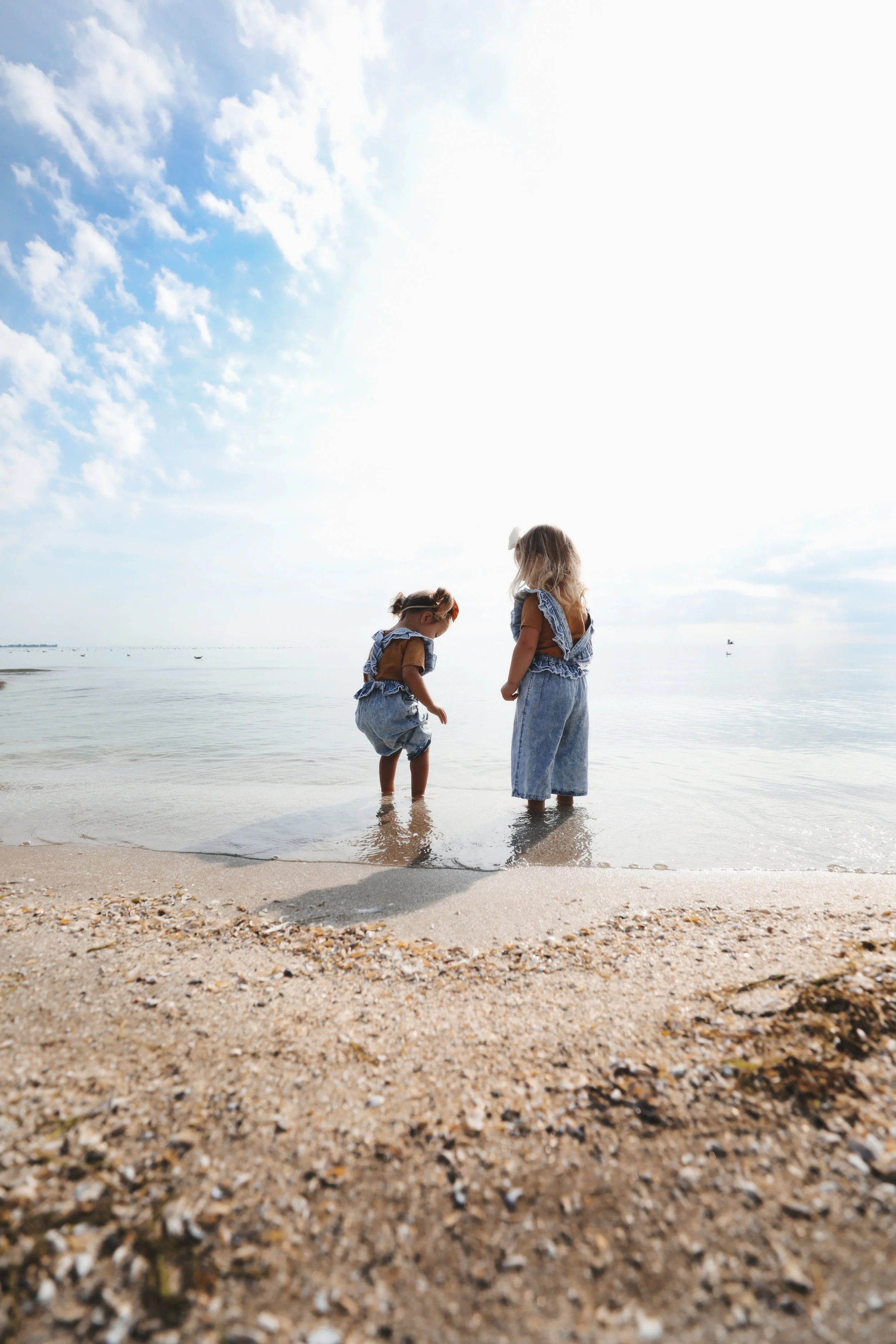 Two young girls with blonde hair, wearing denim clothes, standing in shallow water at the beach during daytime, with ocean and cloudy sky in the background.