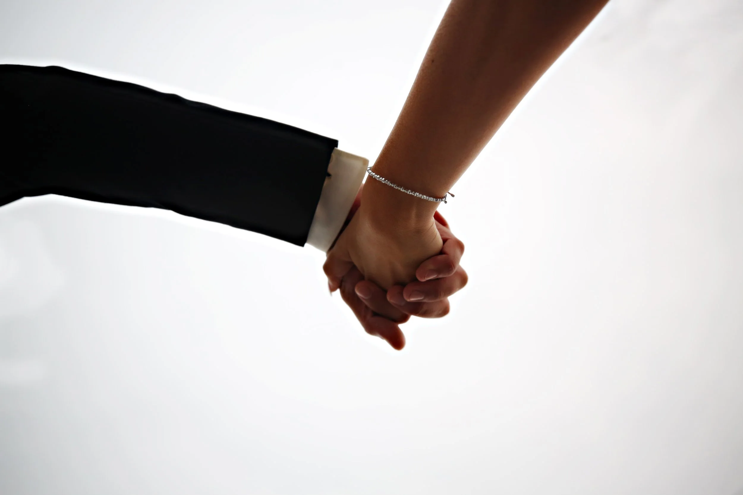 Close-up of two people holding hands against a light background, one wearing a black suit and the other with a bracelet on their wrist.