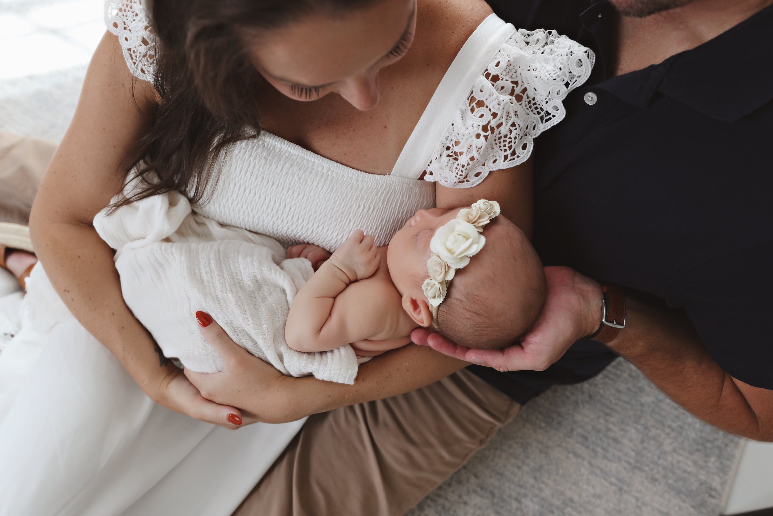 A woman holding a newborn baby girl dressed in a white blanket and wearing a floral headband, while a man supports her head with his hand.