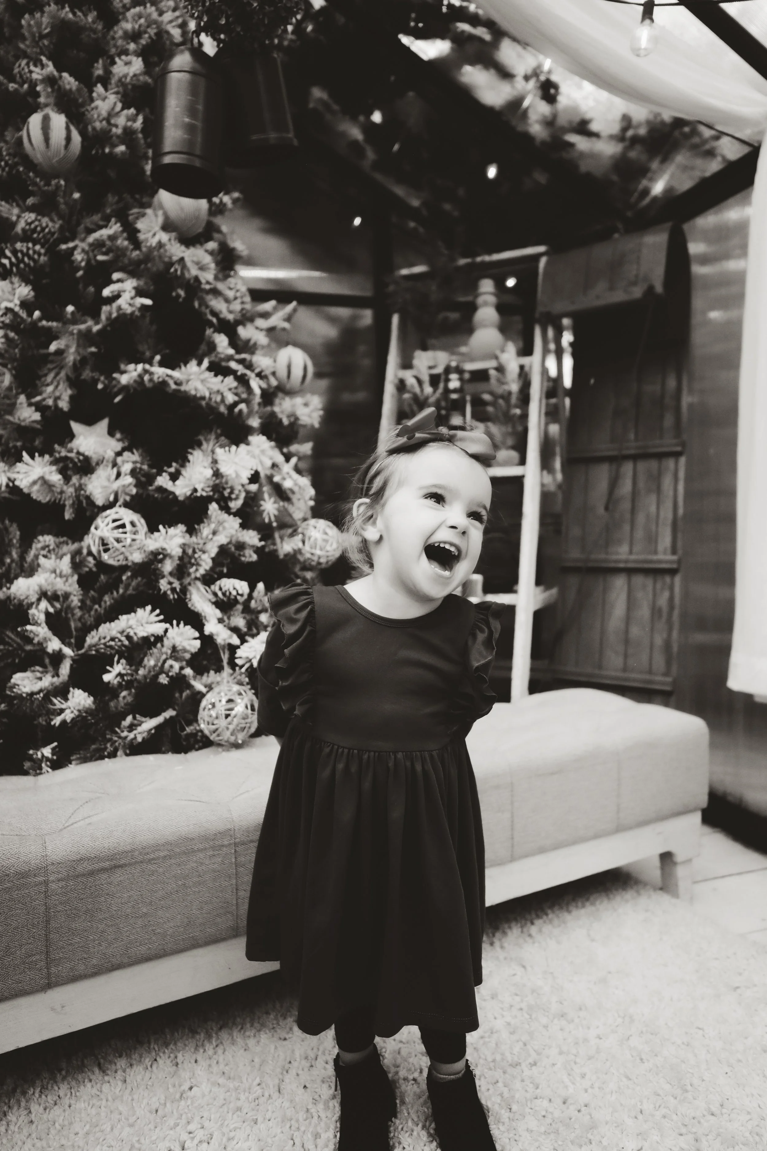 A young girl in a black dress with puffed sleeves and a large bow in her hair is laughing excitedly in front of a decorated Christmas tree inside a wooden-finished room.