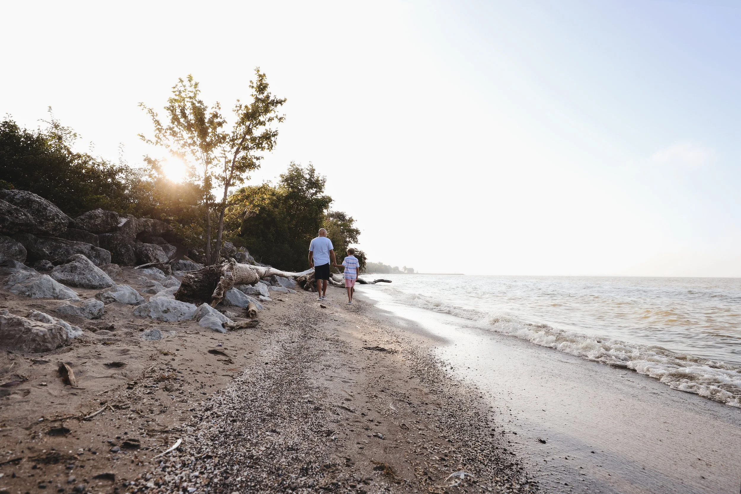 A man and a young girl walking on a sandy beach near the water's edge with trees and rocks nearby, during sunset or sunrise.