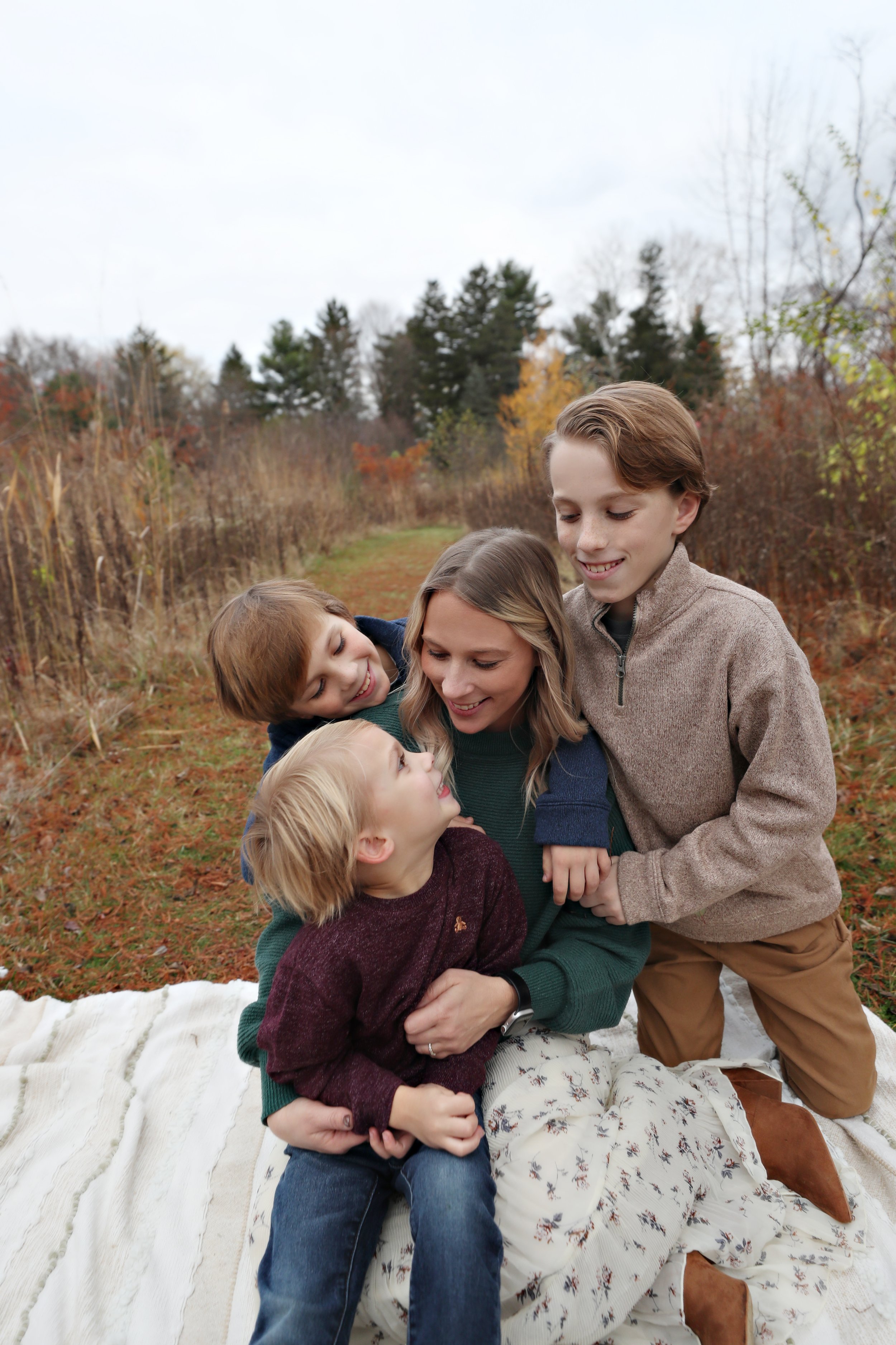 A woman and four children are outdoors on a fall day, sitting on a blanket, smiling and hugging each other.