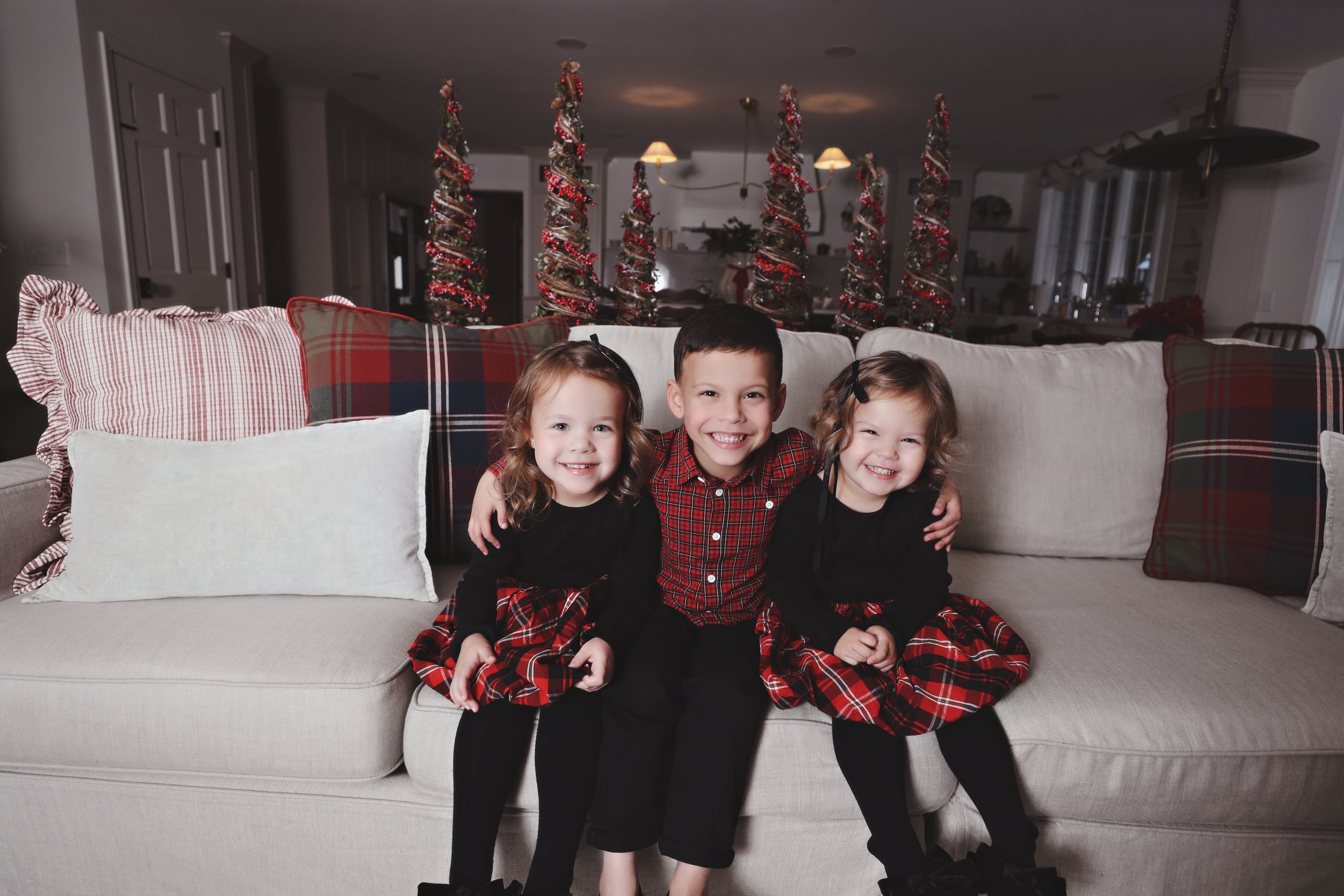Three children sitting on a beige couch, smiling in front of Christmas trees decorated with red ribbons and ornaments, in a festive home setting.