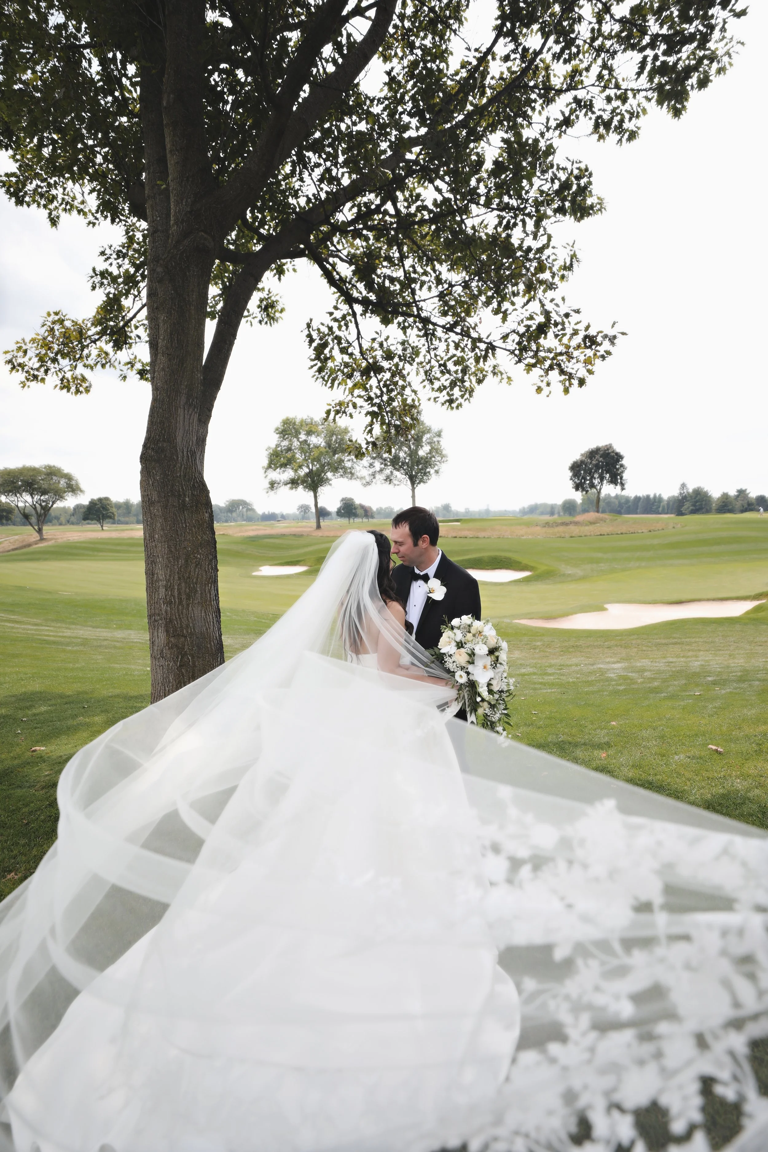 A bride and groom stand close together beneath a large tree on a golf course, with the bride wearing a voluminous white wedding gown and veil, holding a bouquet of white flowers, and the groom in a black tuxedo with a white flower boutonniere.