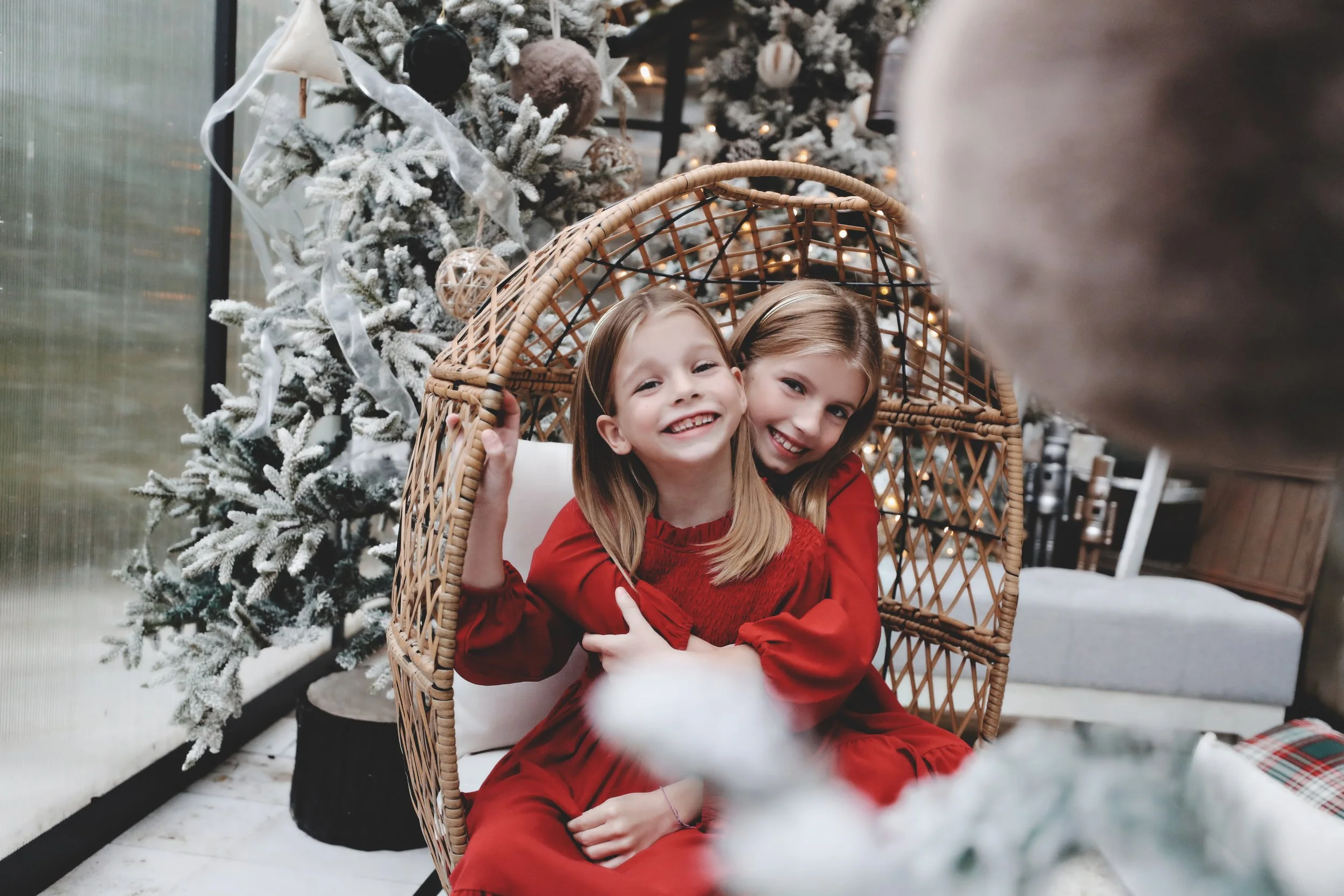 Two young girls in red dresses smiling and sitting in a wicker chair in front of a decorated Christmas tree.