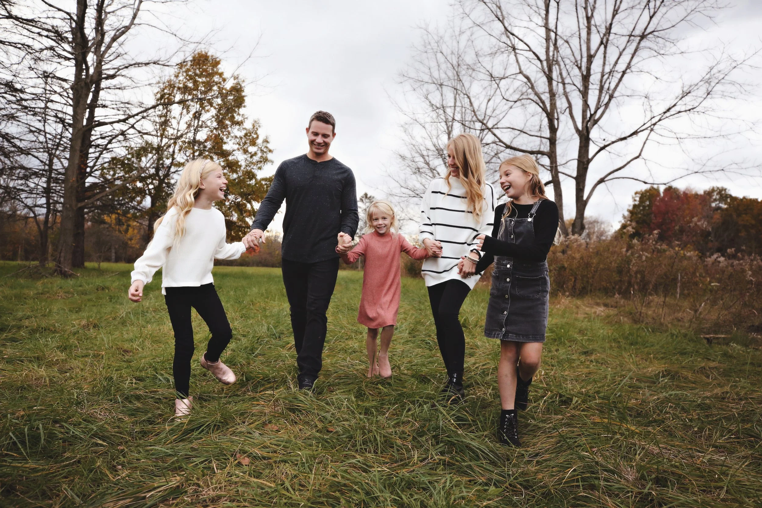 A family of five holding hands and walking in a grassy field during fall.