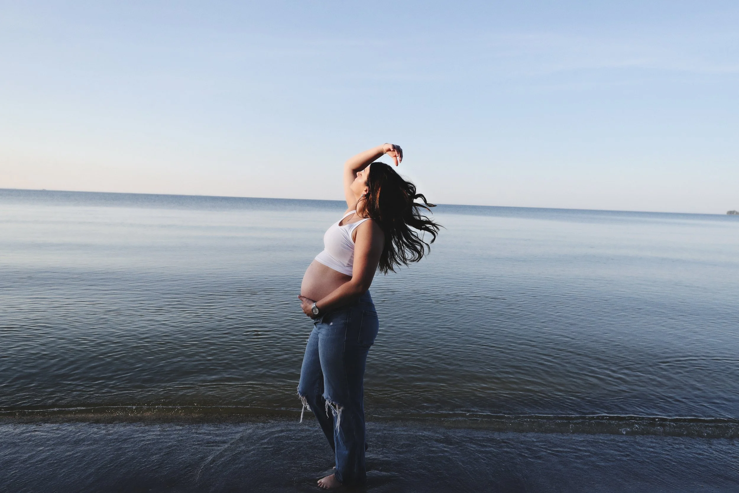 Pregnant woman standing in shallow water at the beach, with long hair flowing, wearing a white top and ripped jeans, holding her belly.