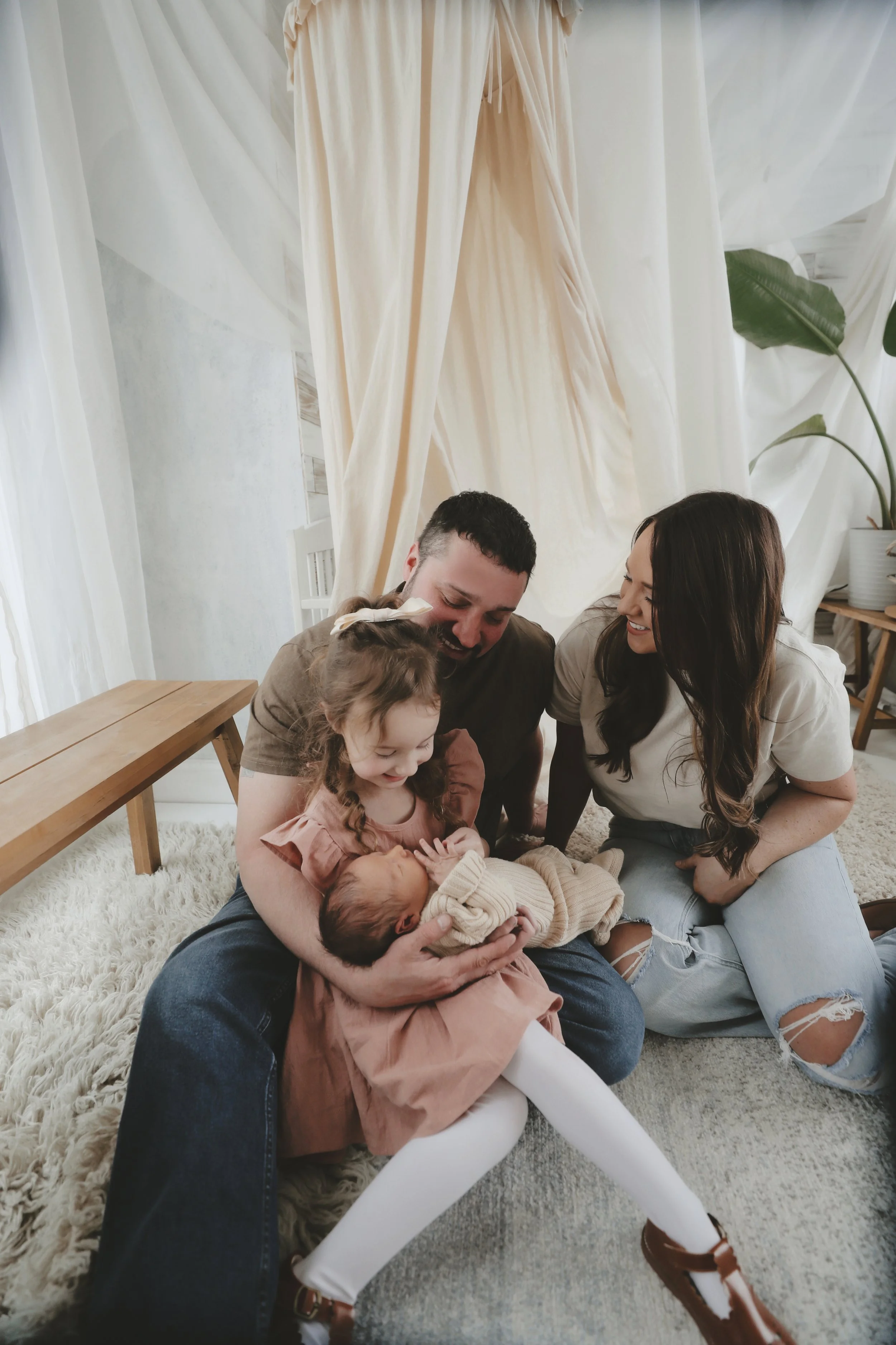 Family of four sitting on a plush rug, smiling and cuddling their two young children, in a cozy, well-lit room with white curtains and a potted plant in the background.