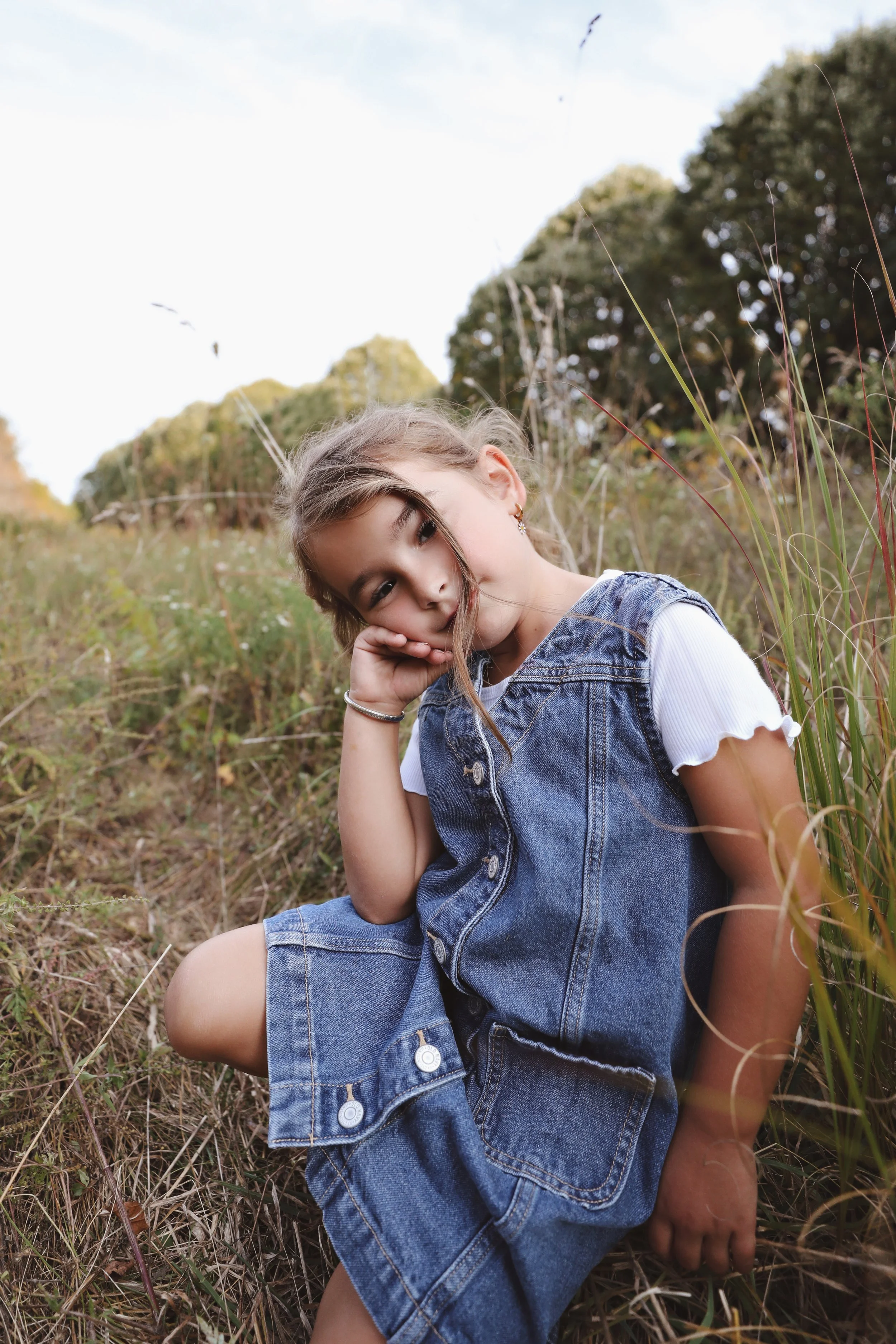 A young girl sitting in a field of tall grass, wearing a denim vest and shorts, looking contemplative with her head resting on her hand.