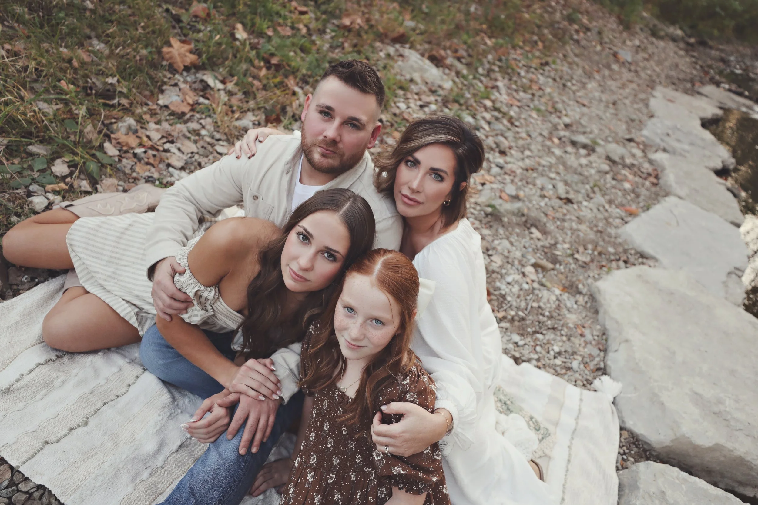 A family of five sitting on a blanket by a rocky riverside. They include a man, woman, and three young girls, all looking at the camera.
