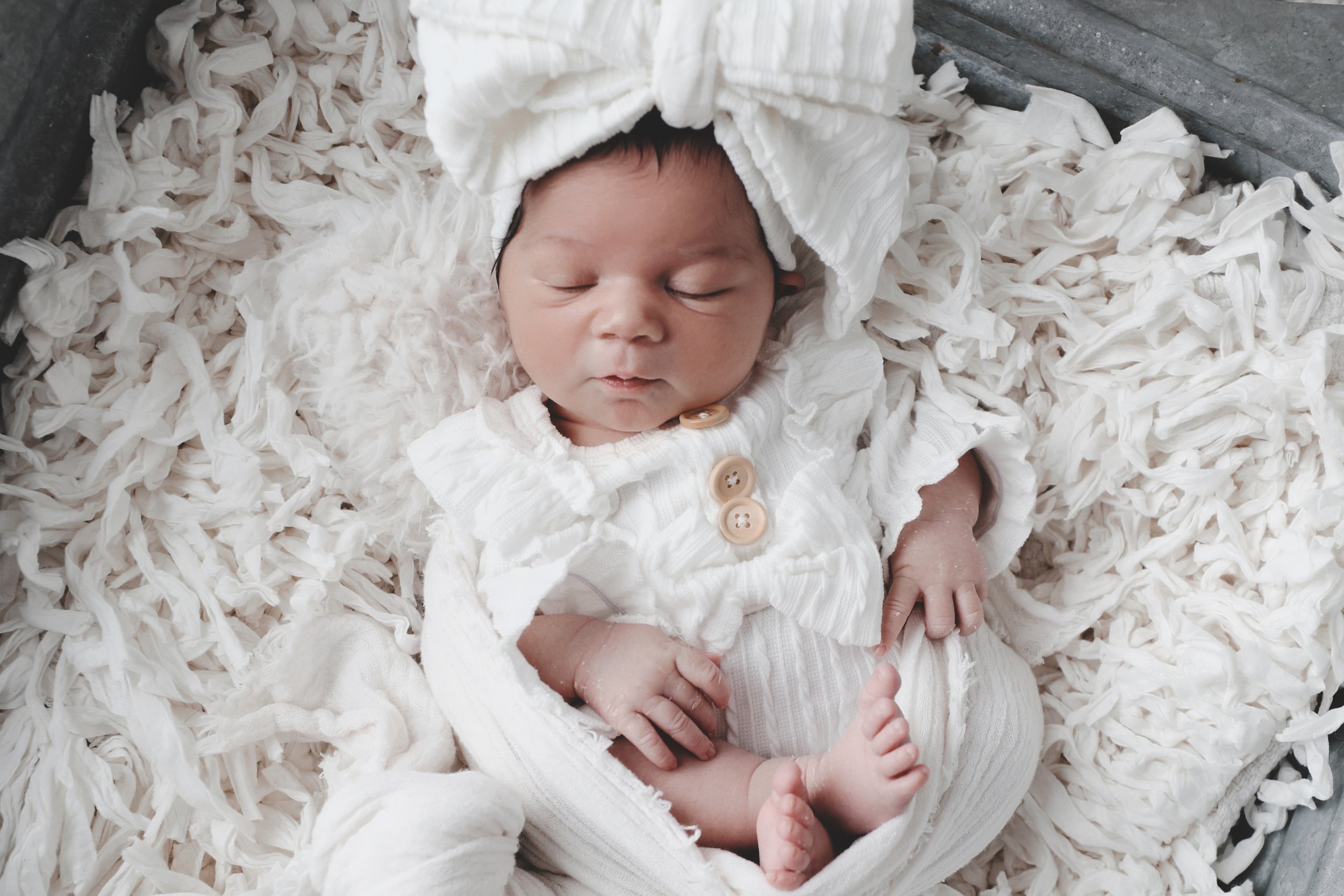 A newborn baby sleeping on a soft, fluffy blanket. The baby is dressed in a white outfit with buttons and a matching hat, with a peaceful expression and tiny hands and feet visible.