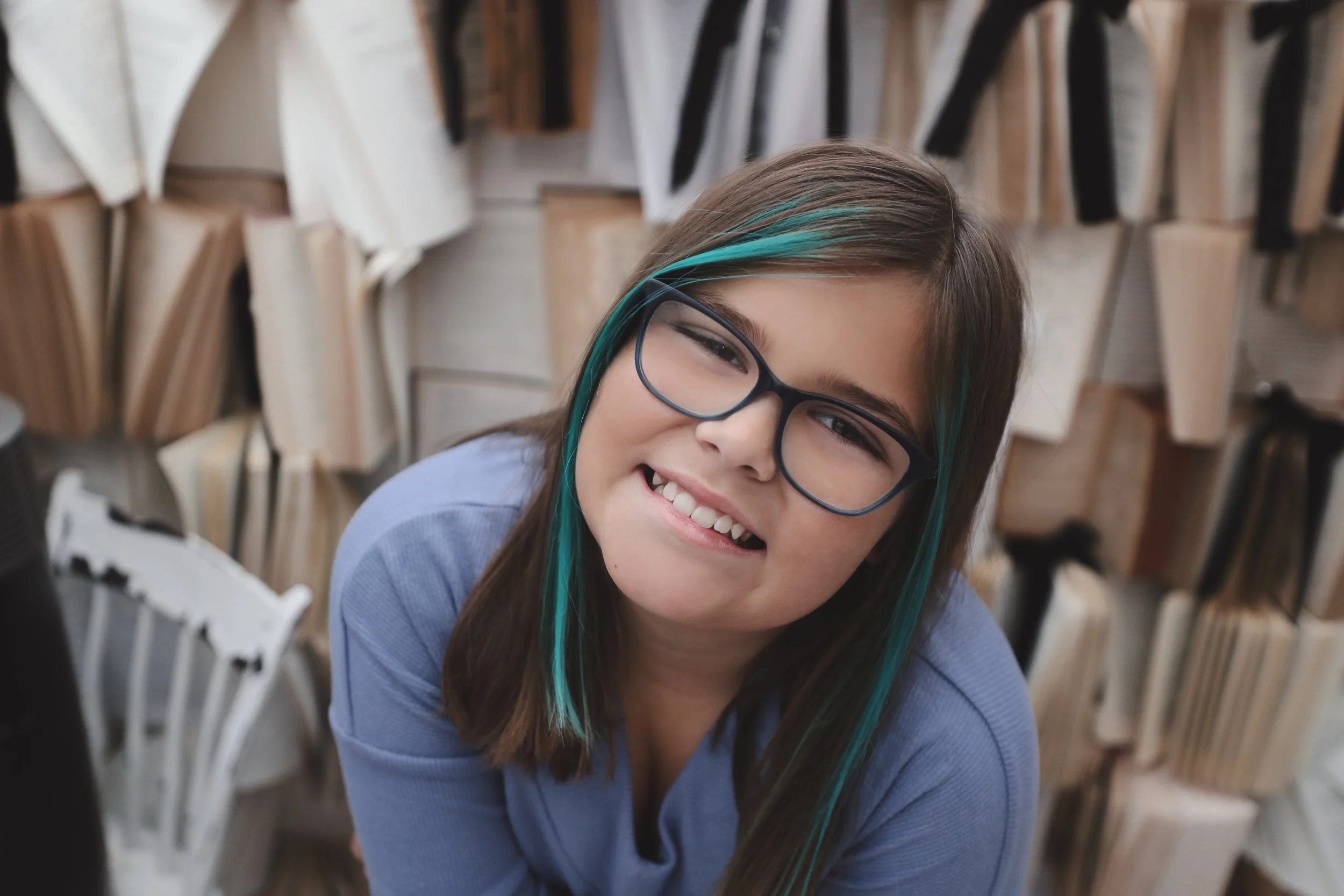 Young girl with glasses and teal streaks in her hair smiling in front of a wall of books.