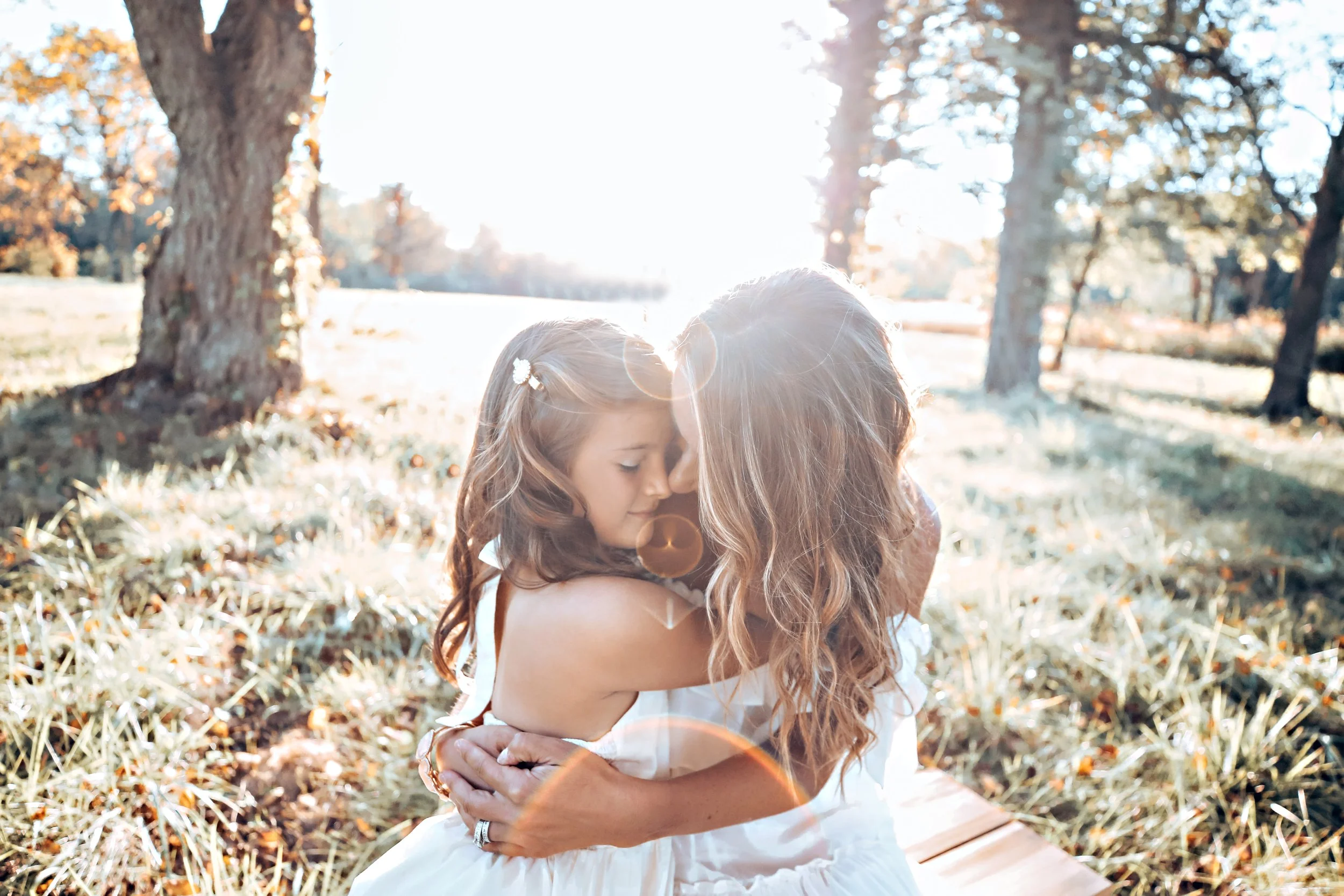 A woman and a young girl sharing a hug outdoors in a sunny park with trees and grass, sunlight creating lens flare.