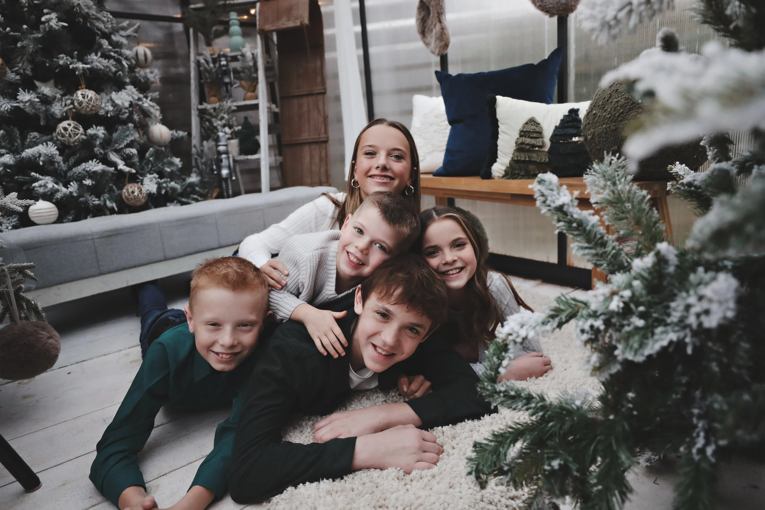 Five children lying on the floor smiling with Christmas trees and holiday decorations in the background.