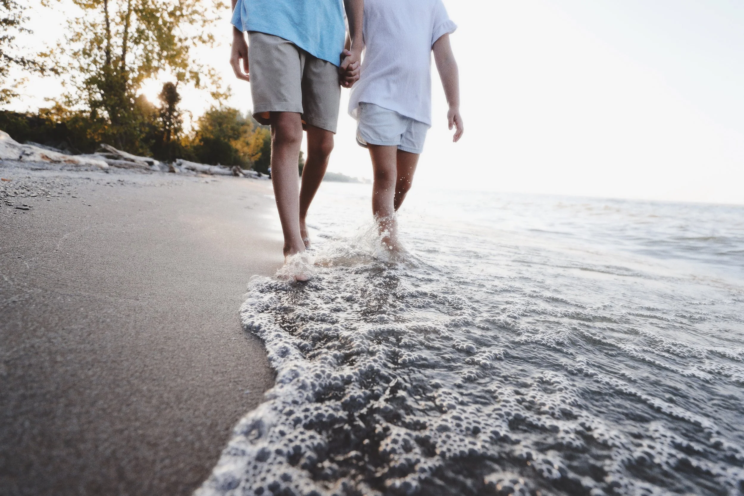 Two children walk hand in hand along the shoreline, with waves touching their feet, on a sunny day.