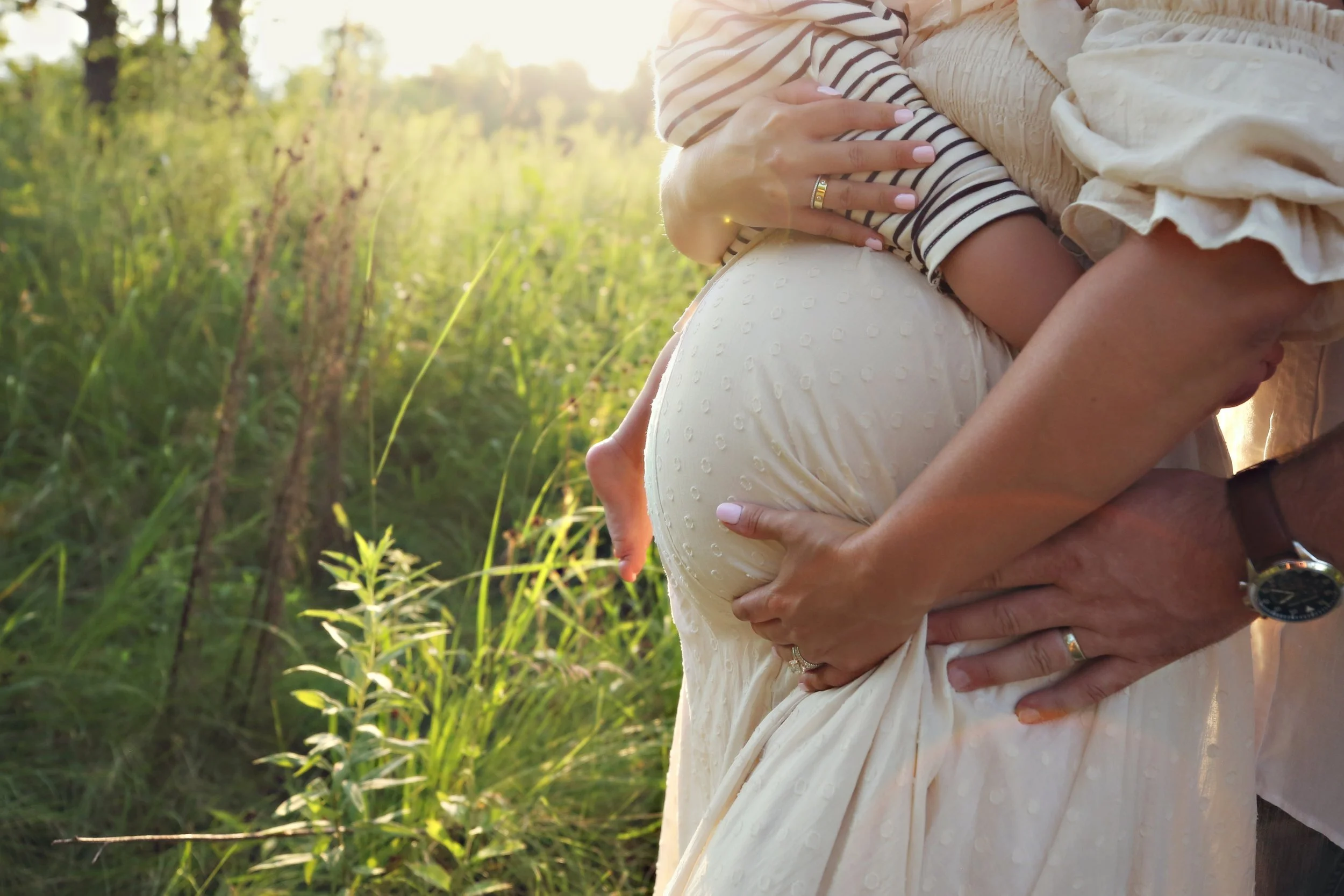 Close-up of a couple embracing outdoors, with a focus on their hands and the woman's pregnant belly, during sunset in a green natural setting.