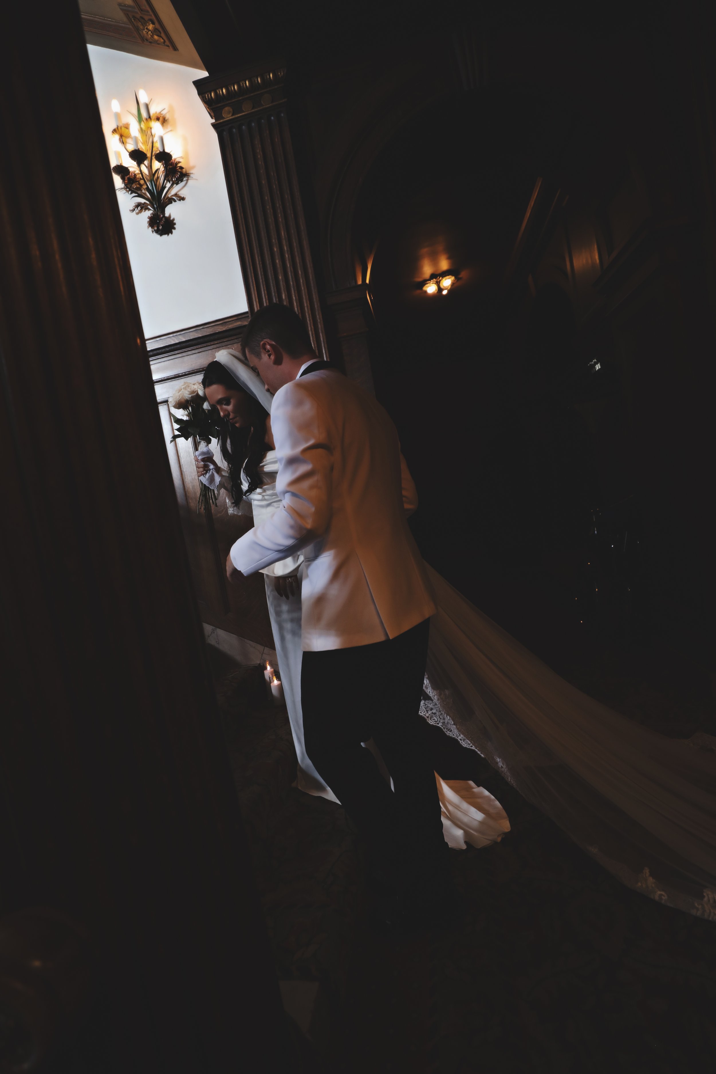 A bride and groom holding hands during their wedding ceremony in a dimly lit room with elegant wood paneling, a floral chandelier, and soft lighting.