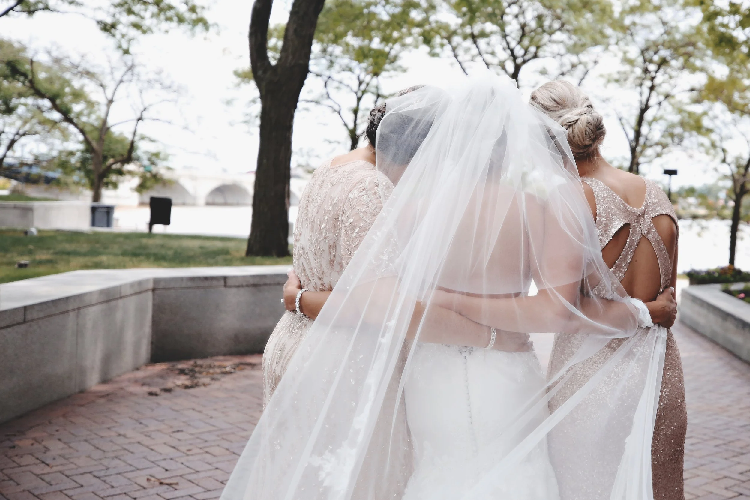 A bride in a white wedding dress and veil embraces two women outdoors near trees and water.