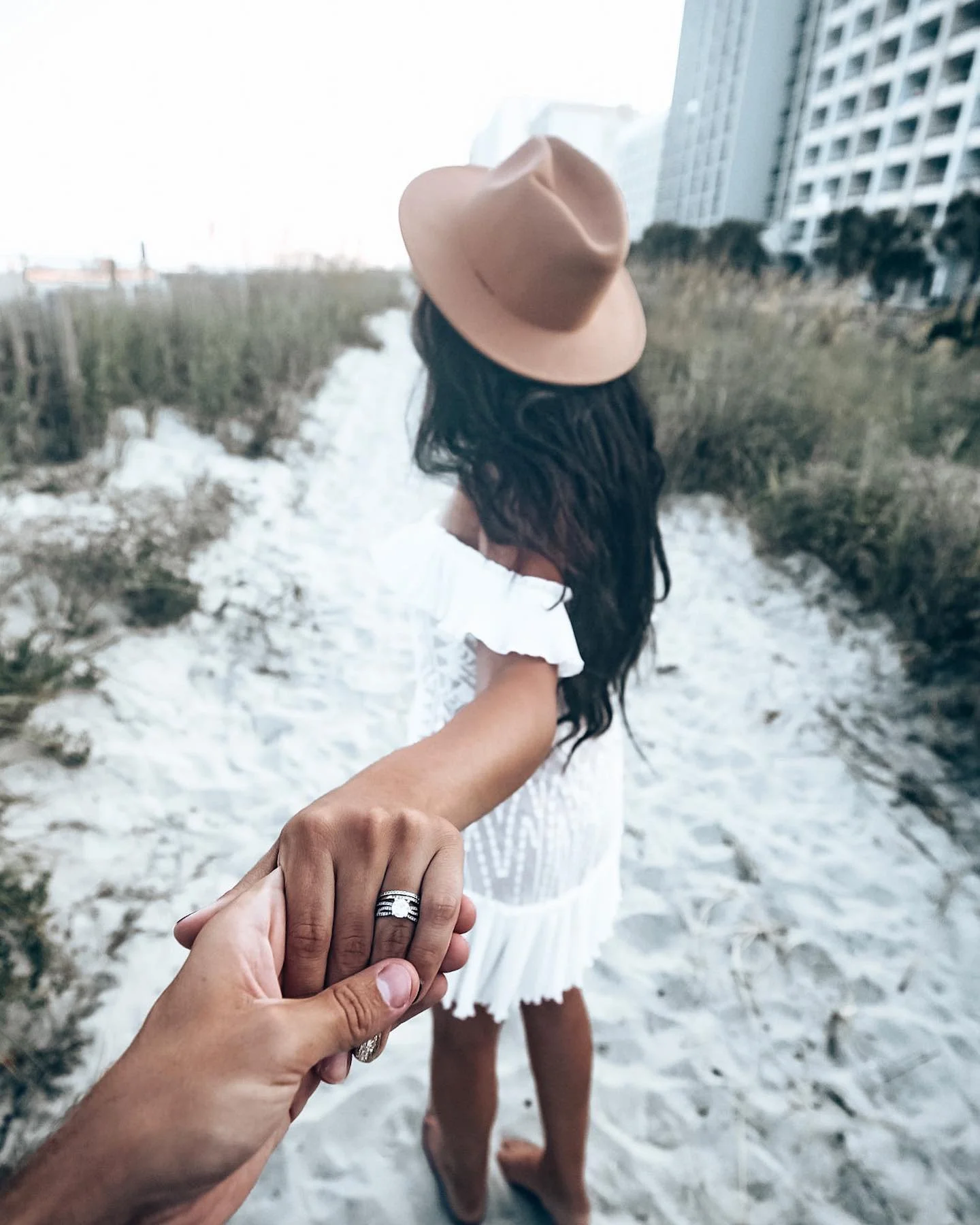 A woman holding hands with a man on a sandy boardwalk, wearing a white dress and a tan hat, with tall buildings and greenery in the background.