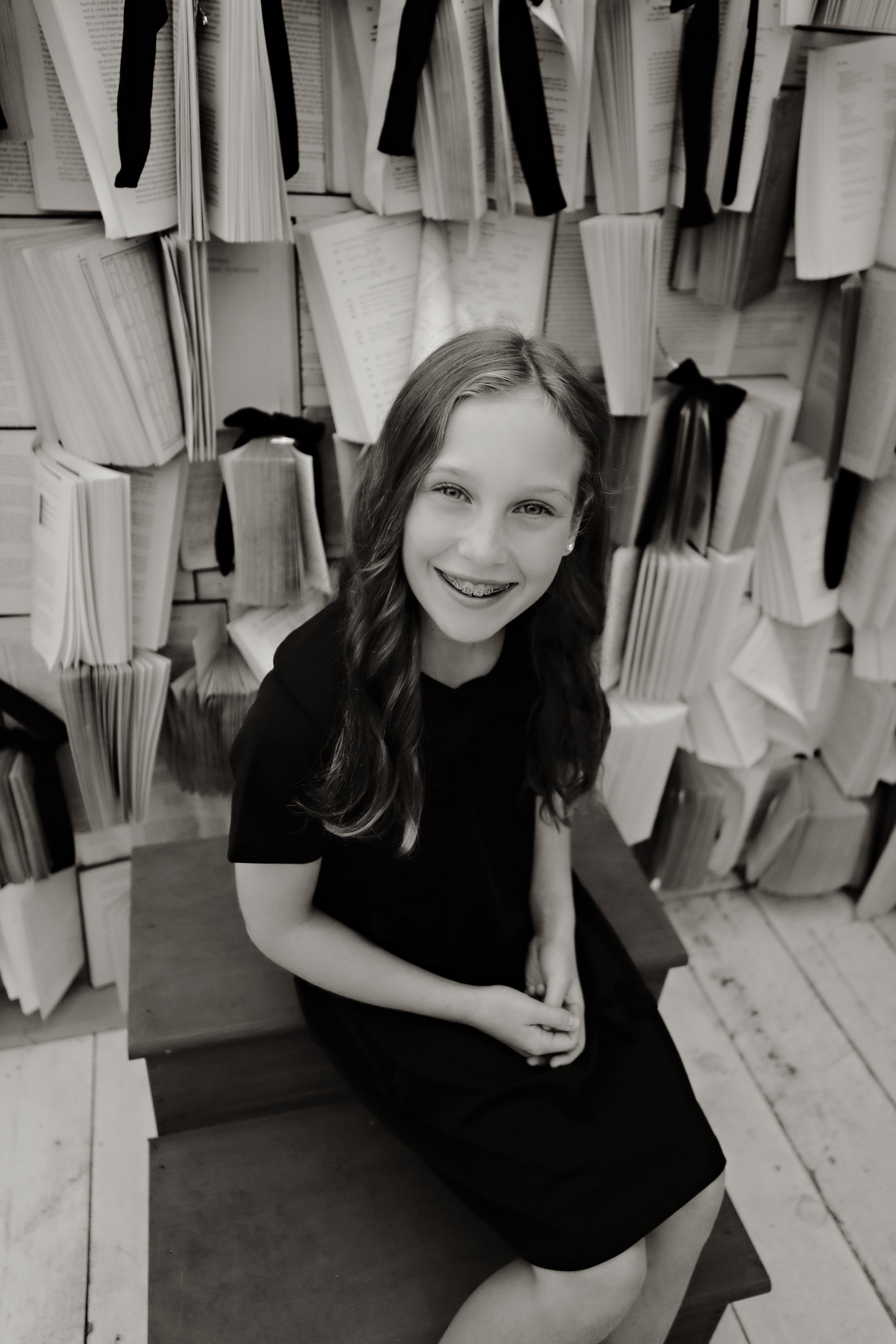 A young girl sitting on a wooden bench in front of a wall covered with open books and black ribbons, smiling at the camera.