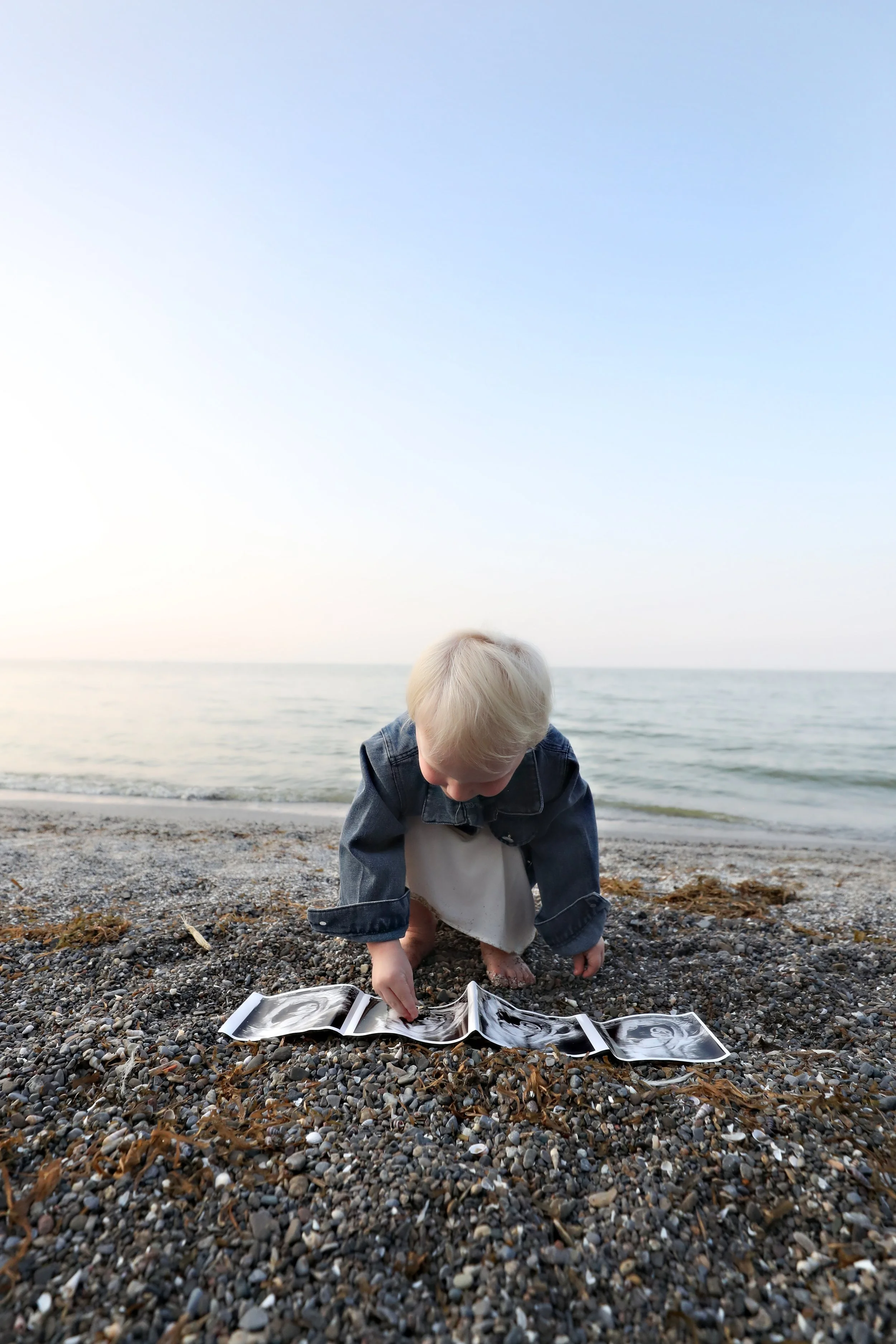 A young child in a denim jacket and white dress kneels on a pebbled beach, looking at and touching an open ultrasound printout laid on the ground with the ocean and sky in the background.