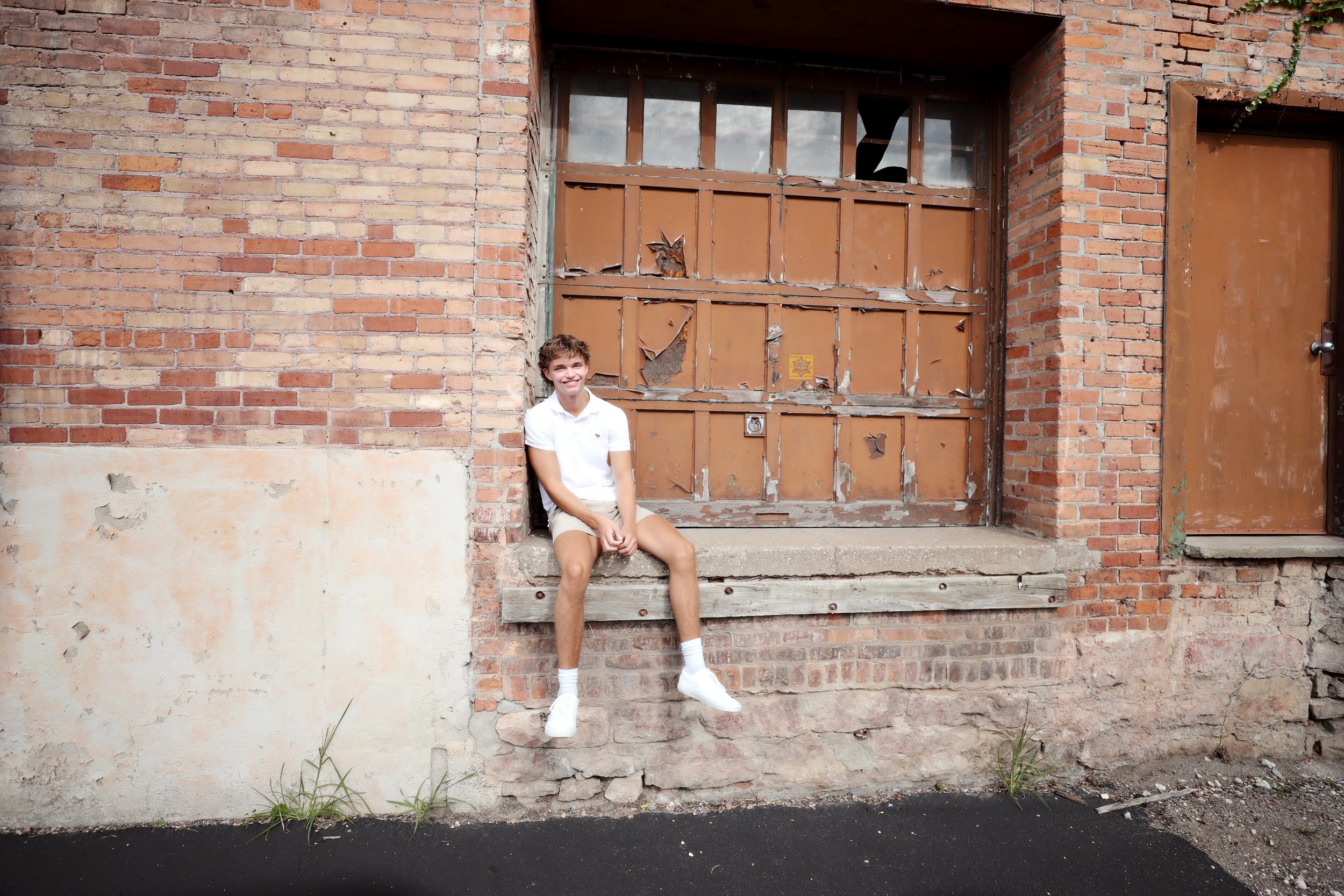 A young man sitting on a wooden ledge in front of a weathered, brown metal garage door on a brick building with broken windows, smiling at the camera.