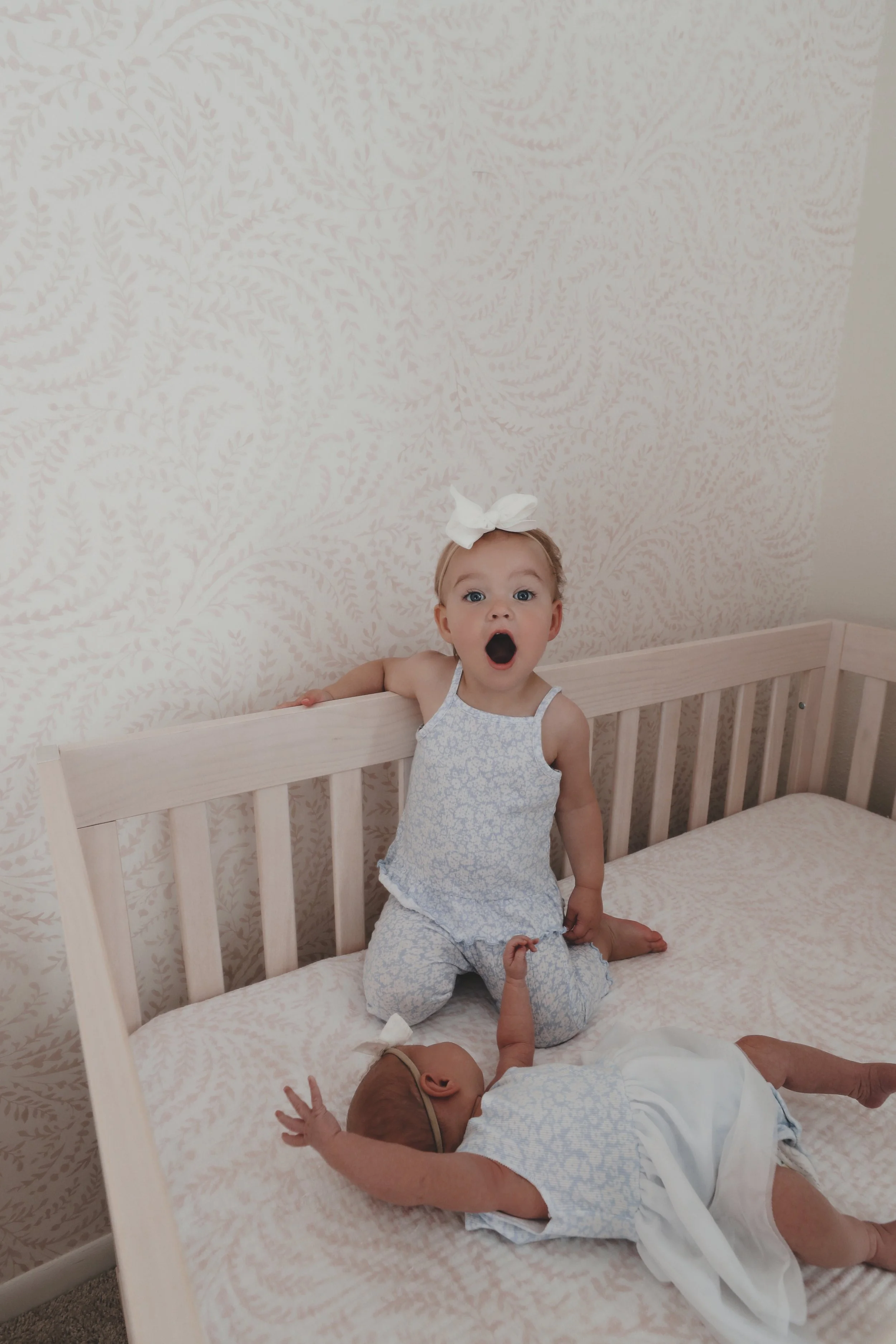 A young girl with a white bow on her head, wearing a blue and white patterned sleeveless top and matching pants, is kneeling on a bed with a surprised expression. A newborn baby lying on the bed is reaching upward with one arm.