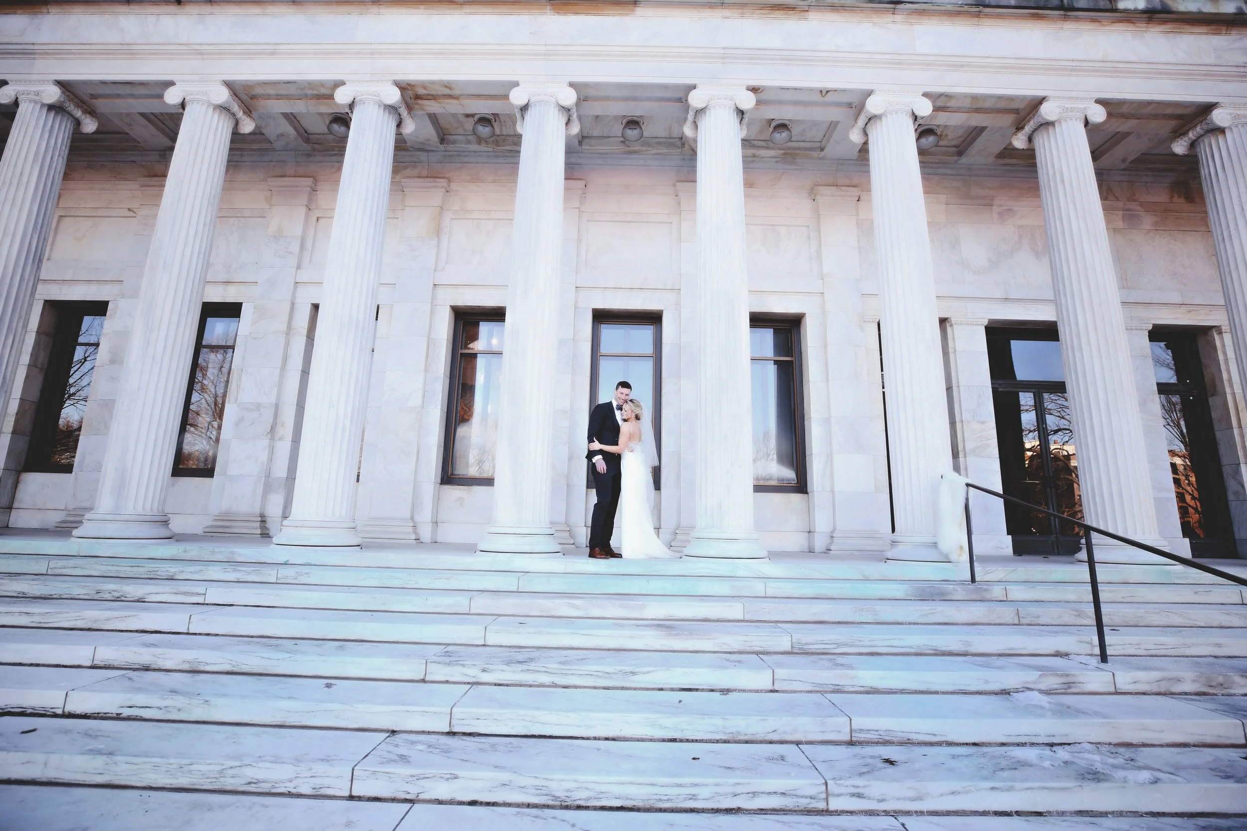 A bride and groom standing on marble steps in front of a classical building with tall columns, during daytime.