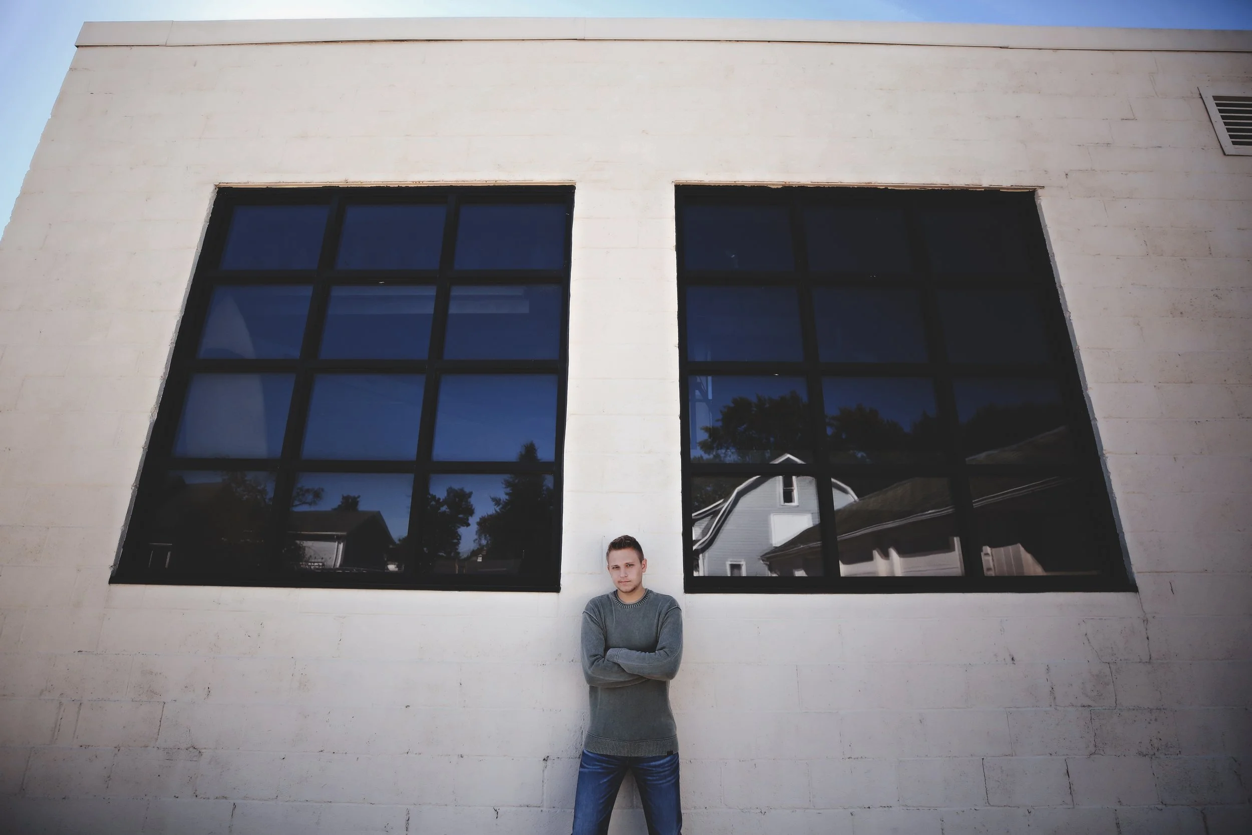 Young man standing with arms crossed in front of a white brick wall with two large black-framed windows reflecting houses and trees, clear blue sky.