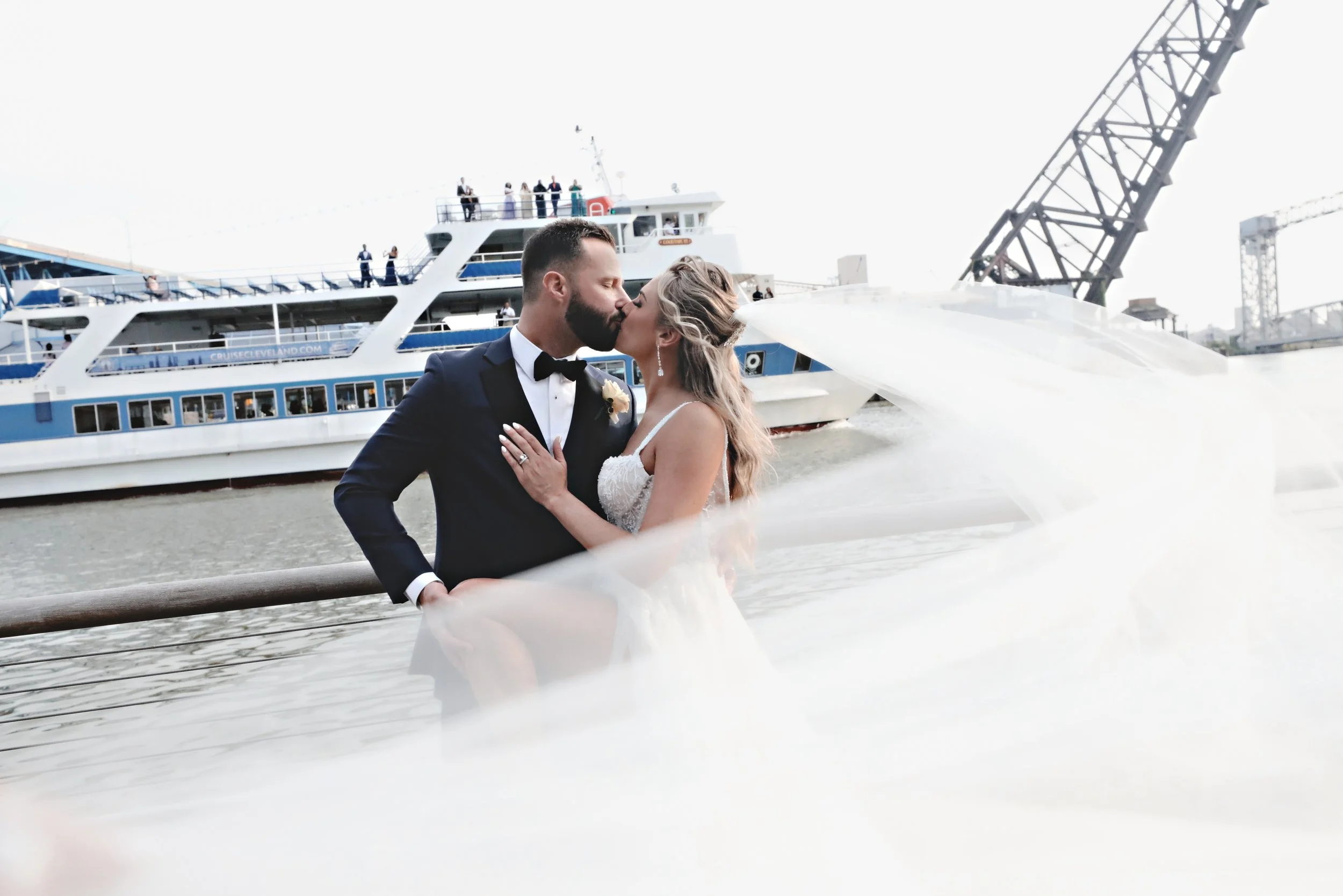 A newlywed couple sharing a kiss by the water with a large cruise ship in the background.