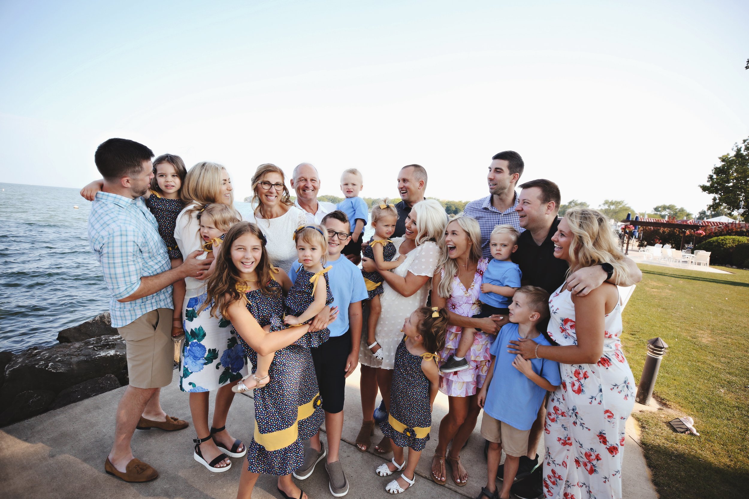 A large family of adults and children smiling and gathered outdoors by the water on a sunny day.