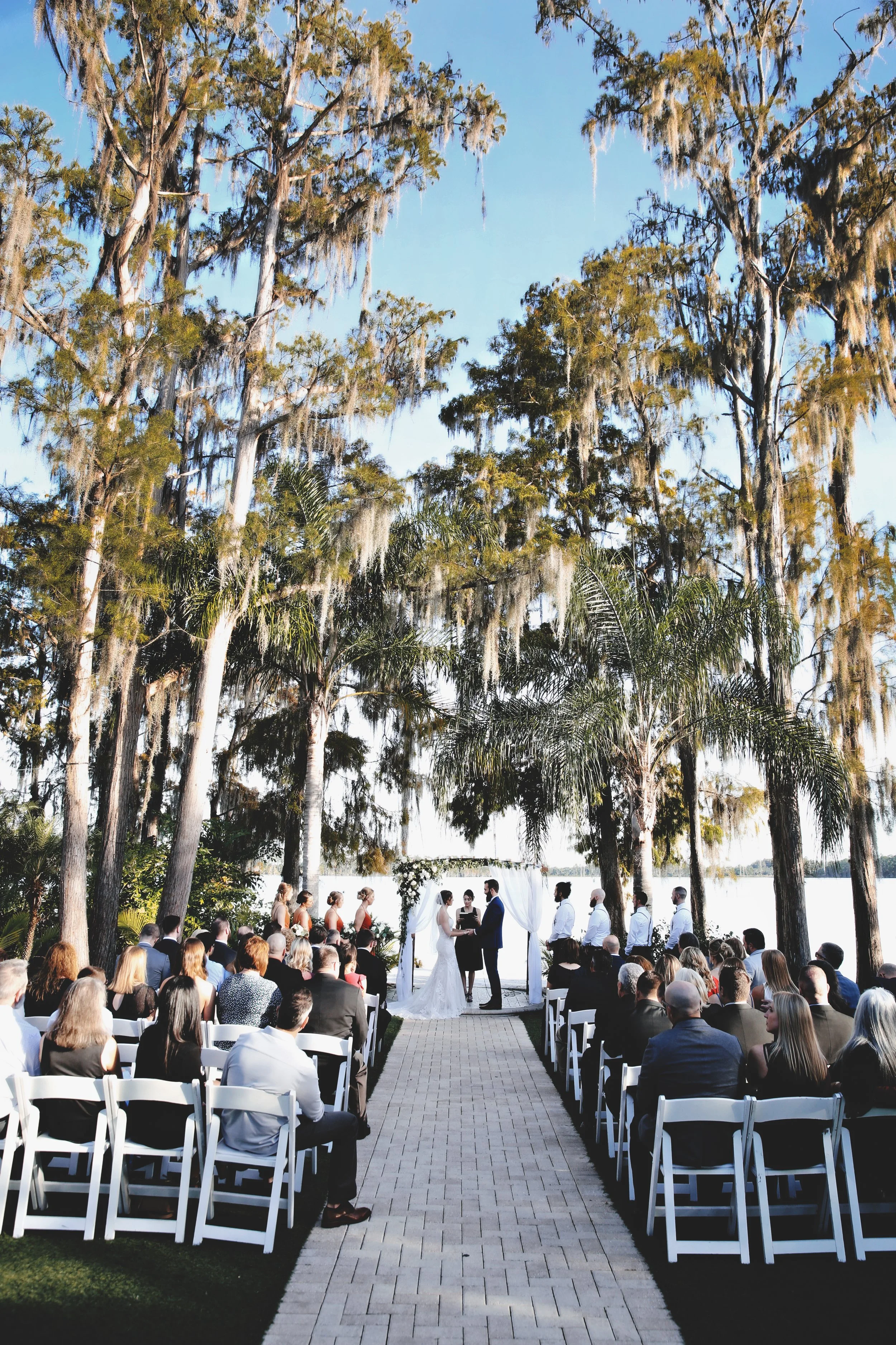 Outdoor wedding ceremony taking place beneath tall trees near a lake, with guests seated in white chairs facing an altar where the bride, groom, and officiant are standing.