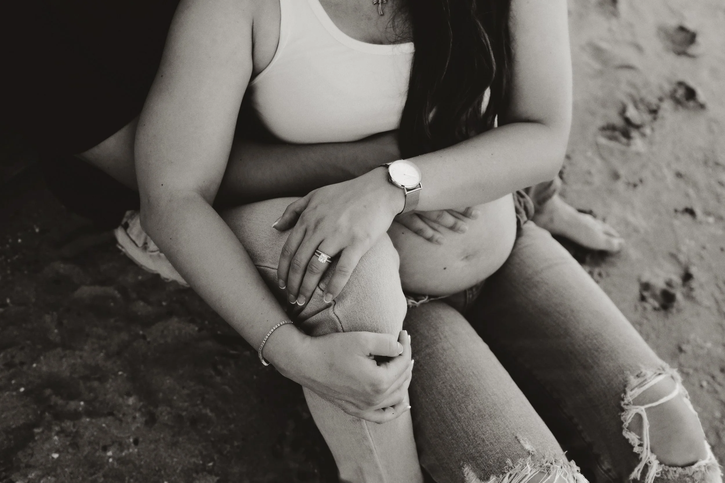 Black-and-white photo of a pregnant woman sitting on the ground with her partner, showing her belly and their intertwined hands.