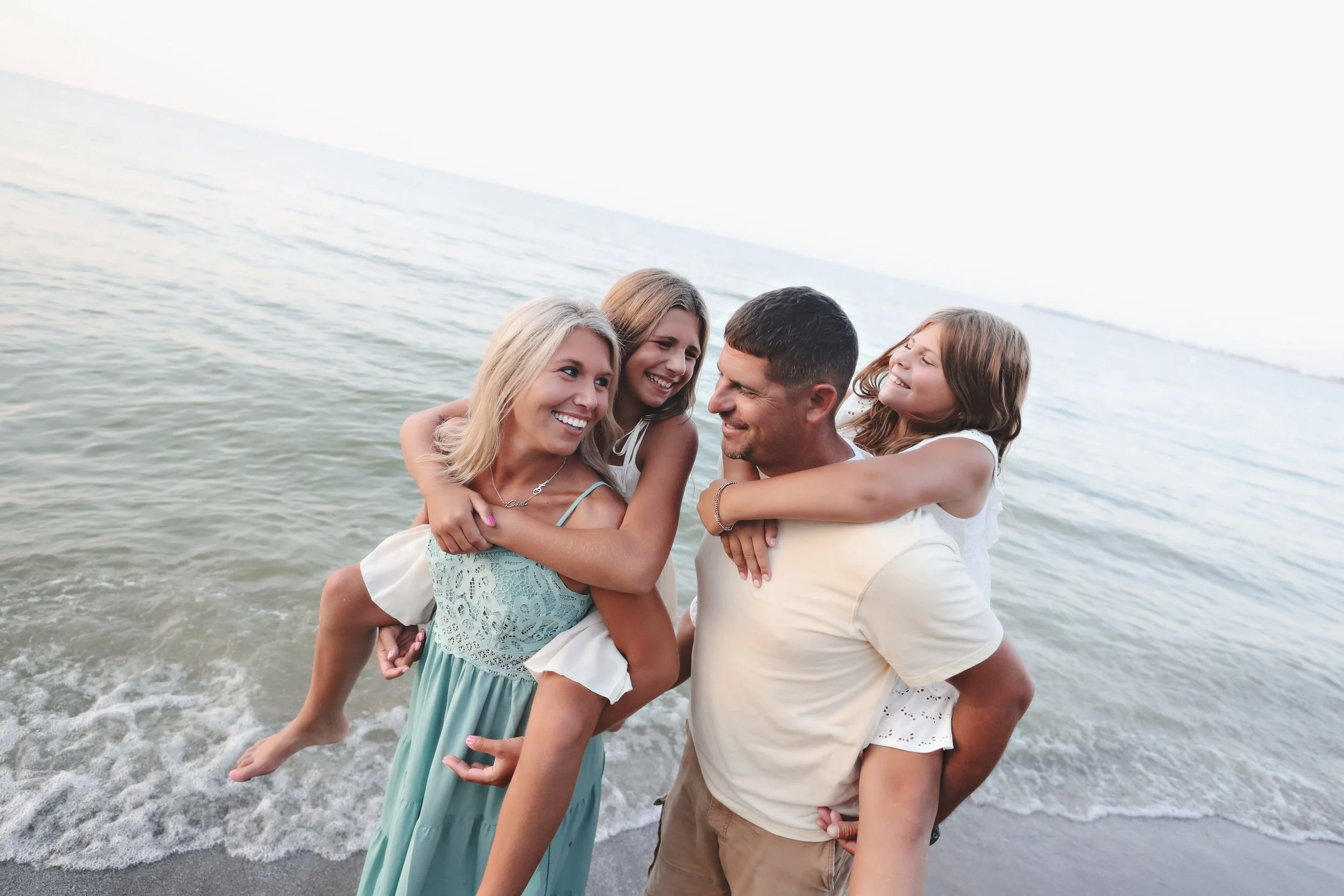 A family of four enjoying a day at the beach, with two children piggybacking on their parents, all smiling and playing by the water.
