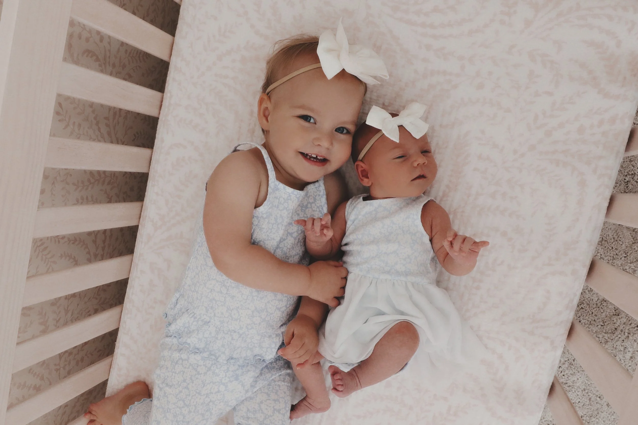 Two young girls lying on a pink bed, one smiling and one looking calm, both wearing white dresses and matching white bows on their heads.