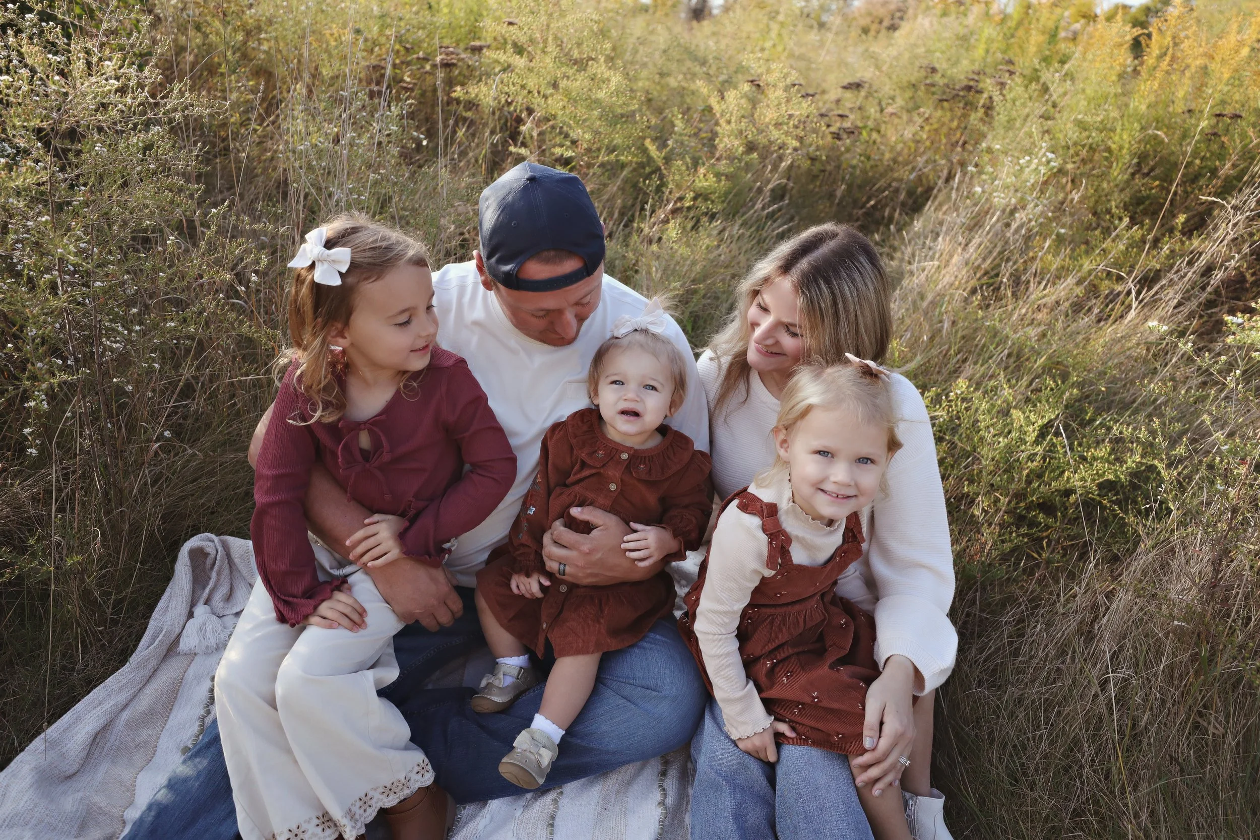 A family of six sitting on a blanket in a grassy outdoor area during fall, with three young girls, one man, and one woman smiling and sitting close together.