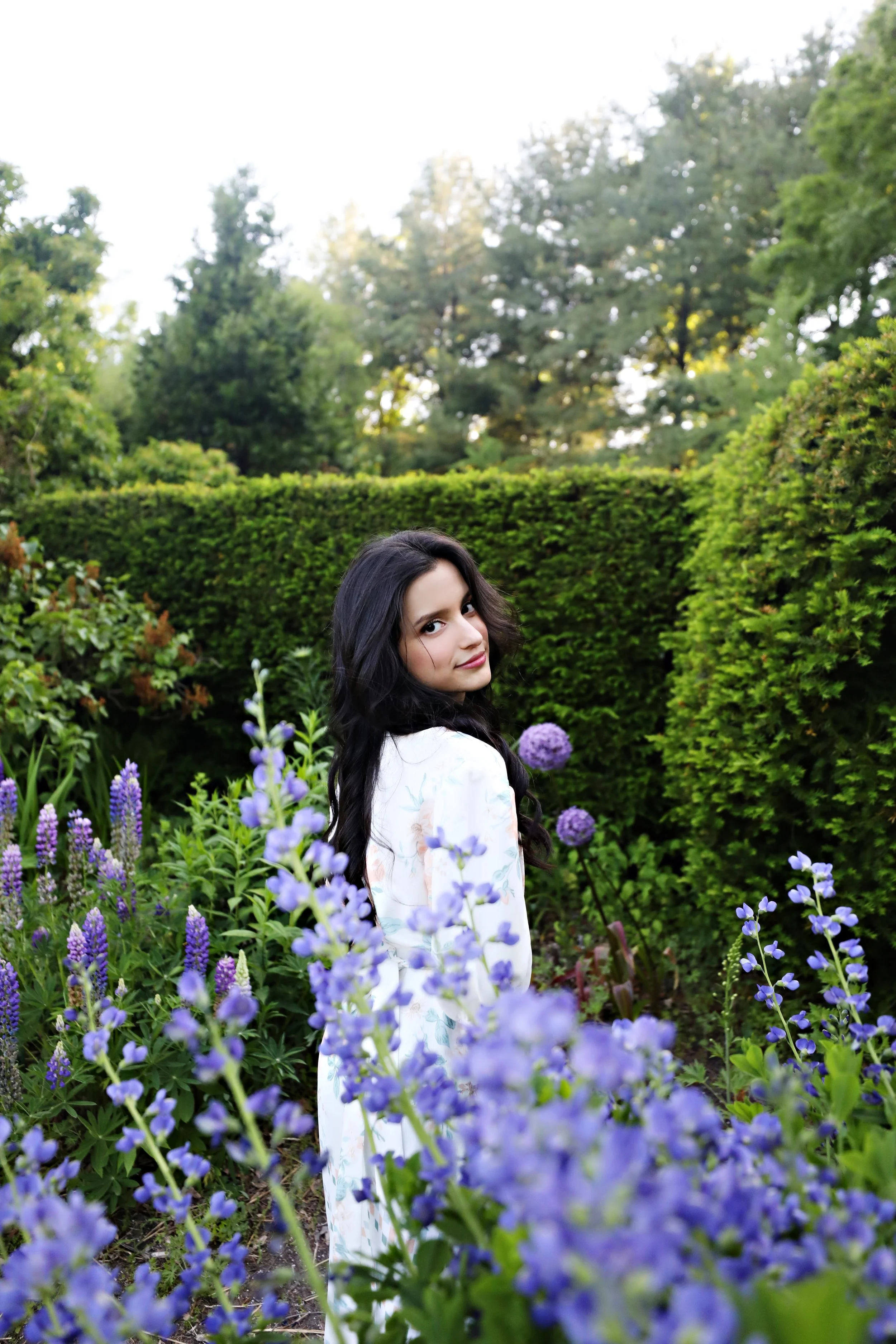 Young woman with long dark hair looking back in a garden with purple flowers and green hedges.