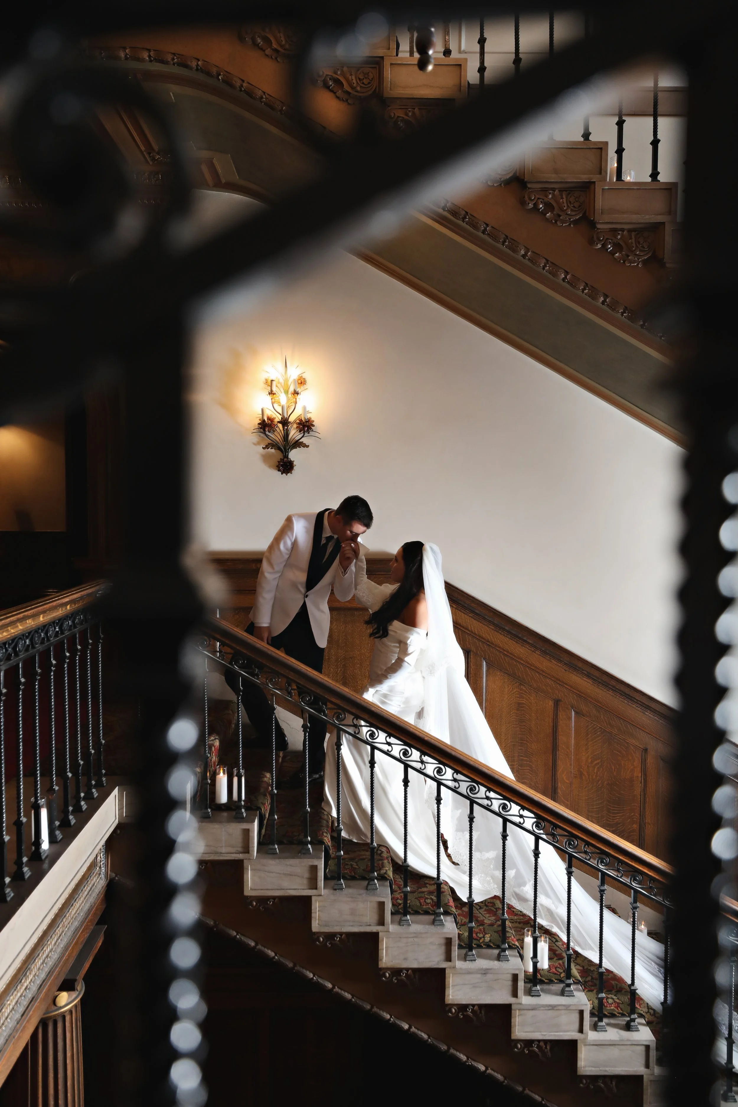 A bride and groom standing on a staircase, with the groom kissing the bride's hand, seen through a decorative iron railing in an elegant, wood-paneled interior with a wall sconce light.