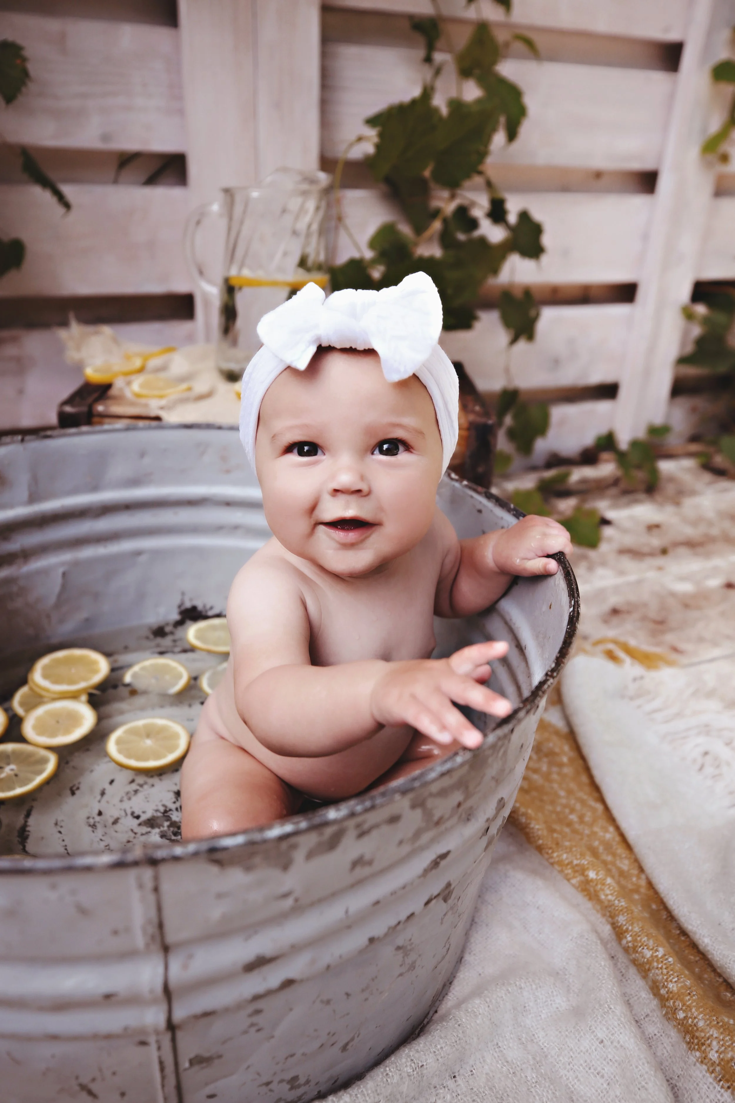 A baby with a white bow headband sitting in a metal tub filled with lemon slices, outdoors with a wooden fence and greenery in the background.