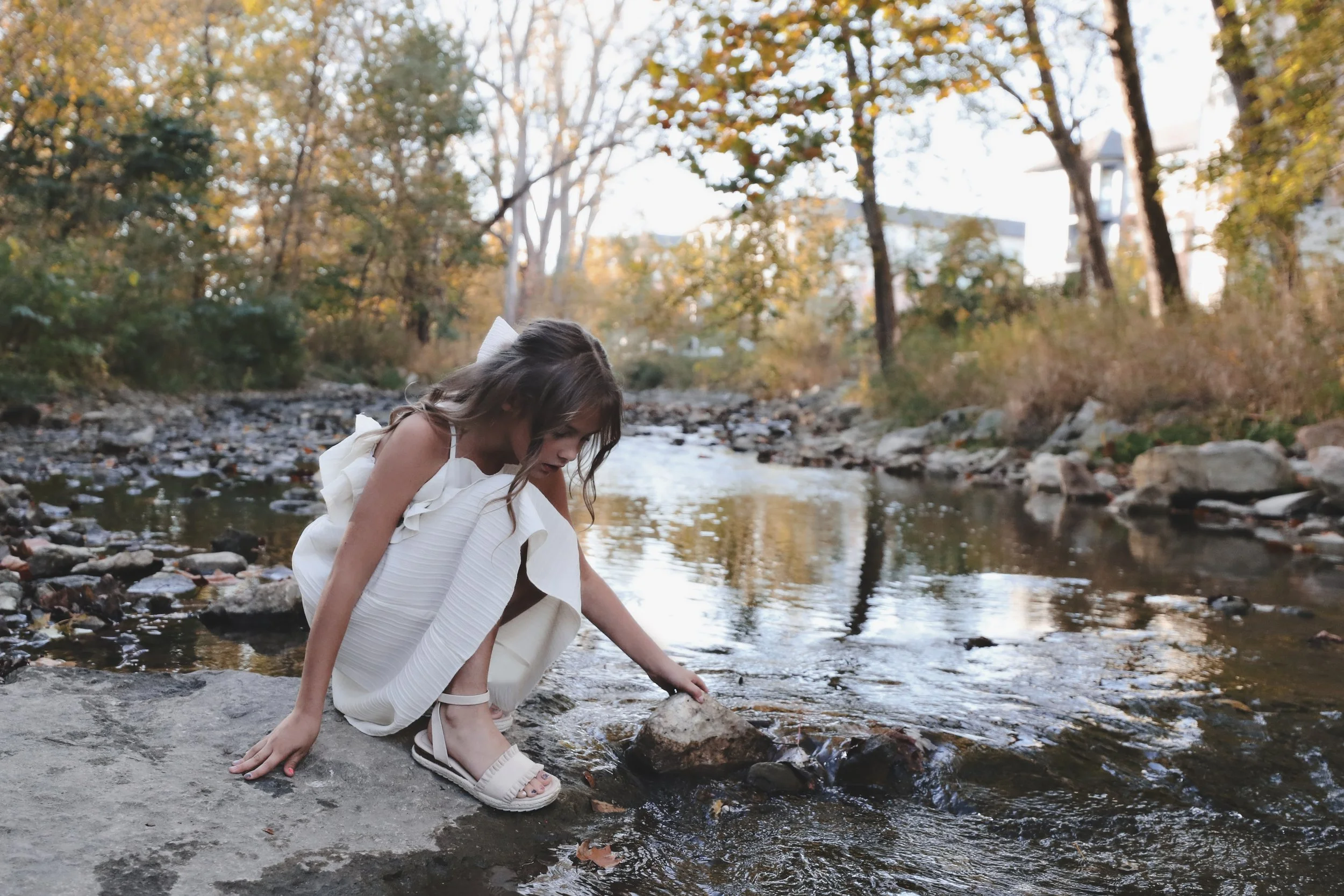 A young girl in a white dress and sandals kneels by a creek, reaching into the water surrounded by trees with fall foliage.