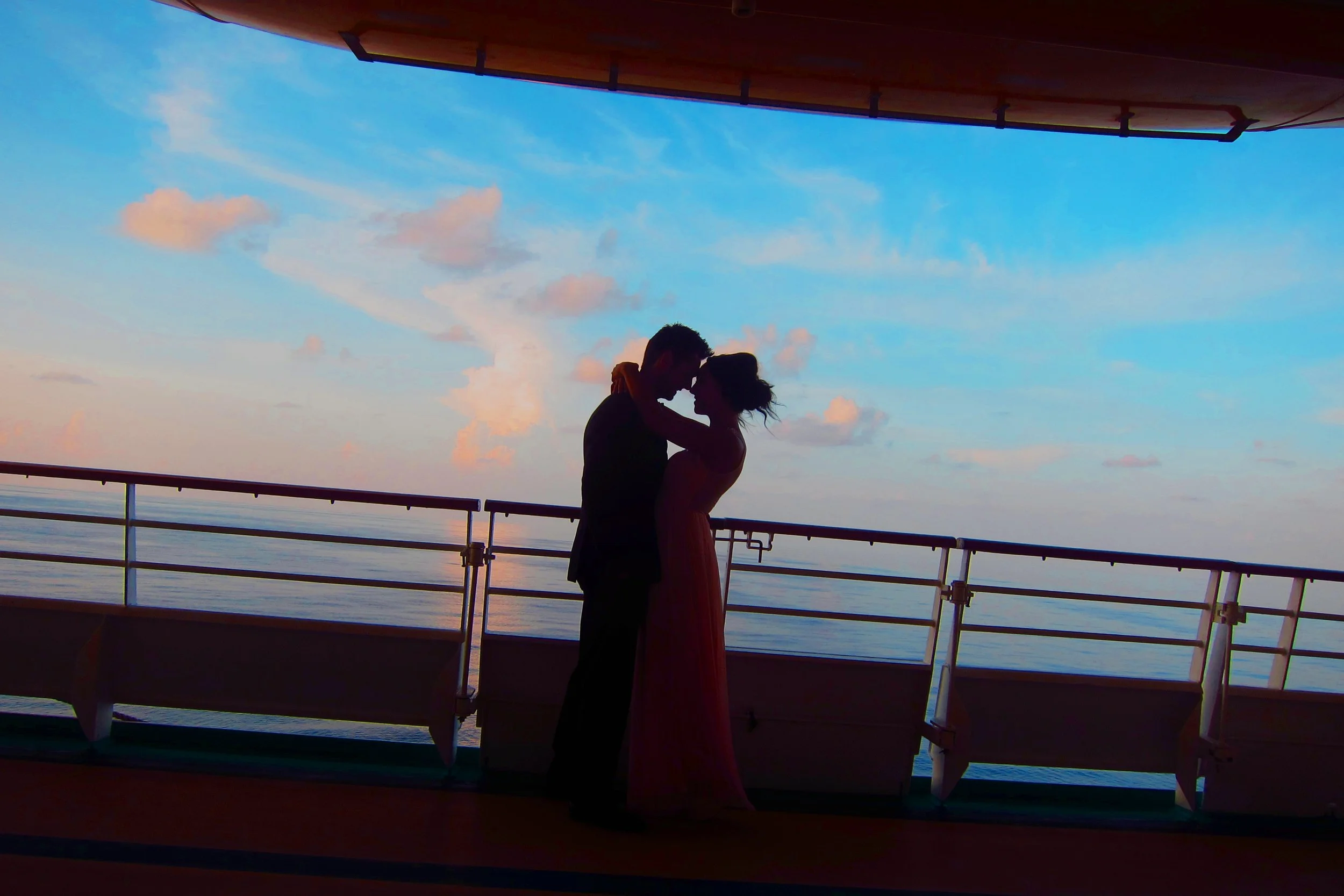 Silhouetted couple embracing on a ship deck at sunset, ocean and sky with clouds in background.