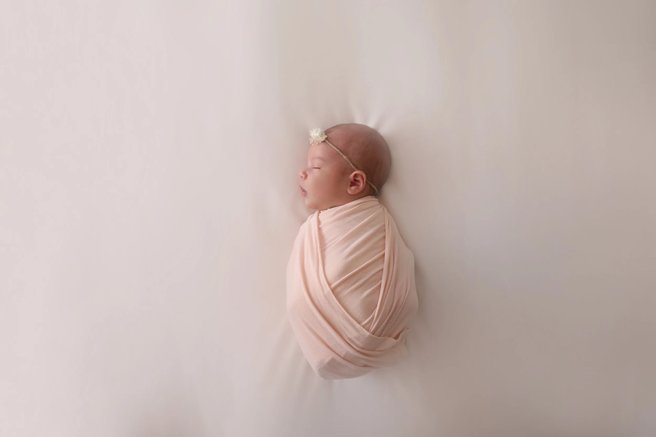 A newborn baby sleeping, wrapped in a pink blanket, with a small floral headband, lying on a white surface.