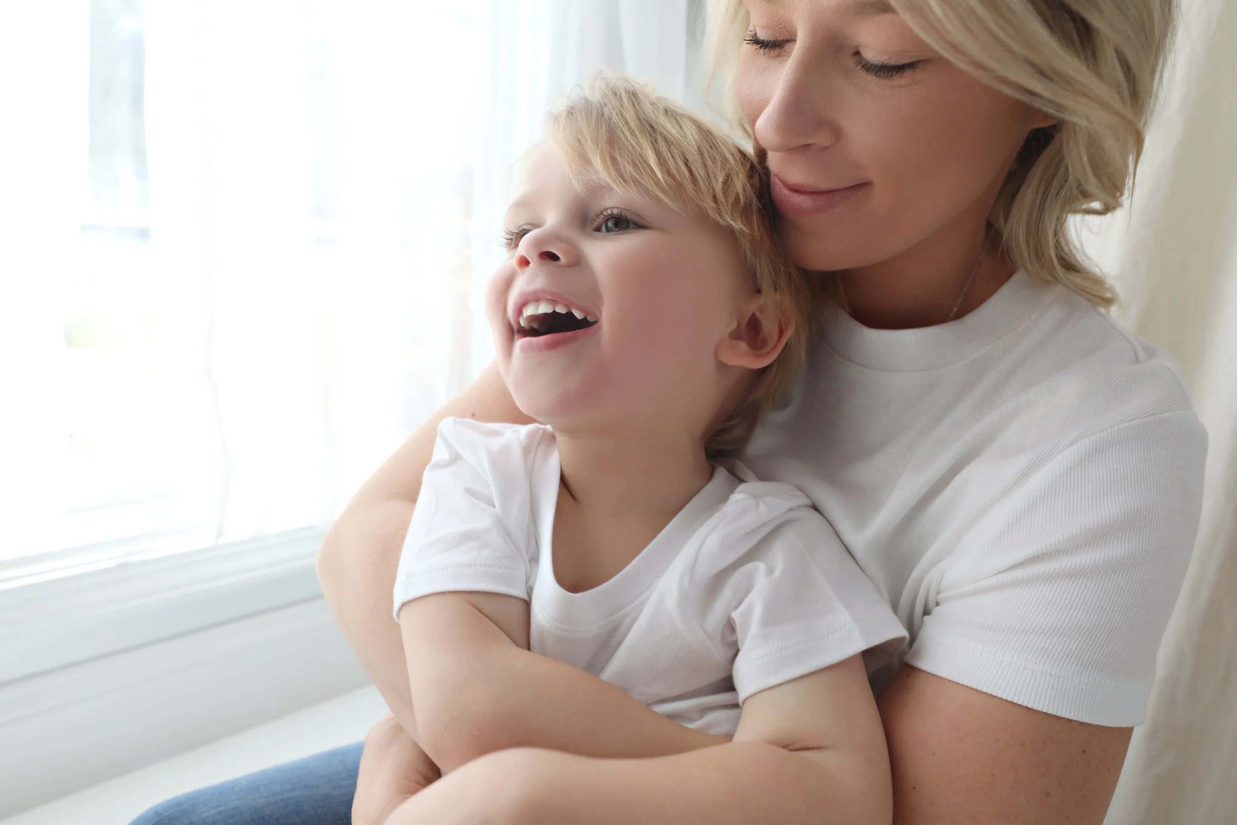 A woman happily hugging a smiling young boy near a window.